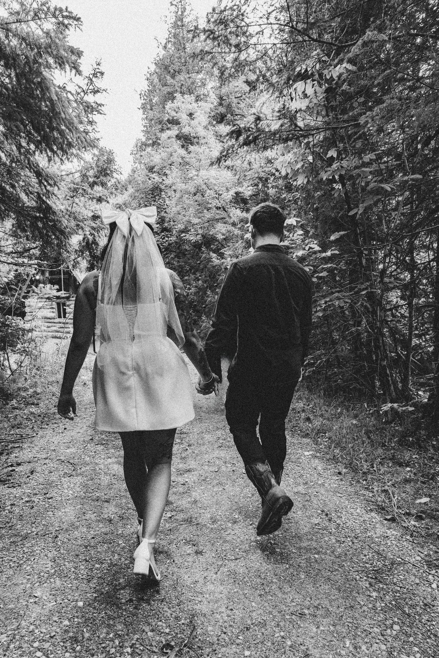 Black and white photo of bride and groom walking down a forest path holding hands.