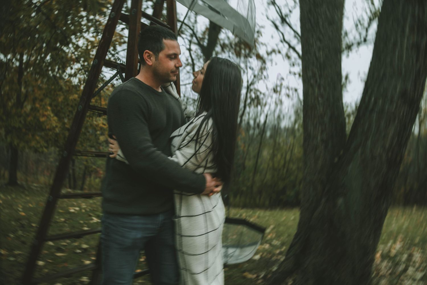 A couple embraces near trees in an outdoor autumn setting during an engagement photo session.