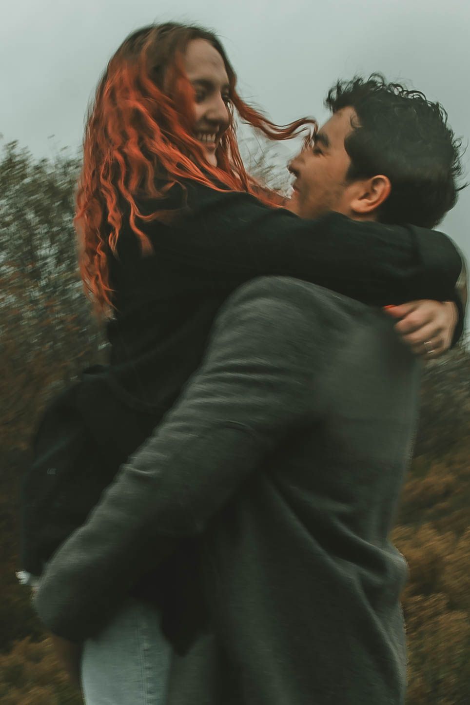 A couple in dark clothing embraces passionately outdoors with red hair flowing in the wind.