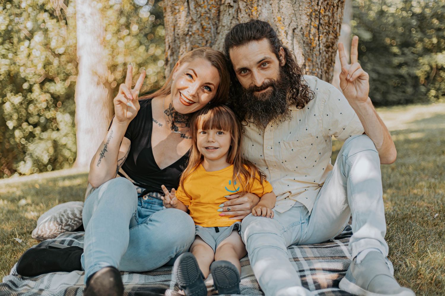 A family of three sits together outdoors making peace signs while smiling in warm sunlight.