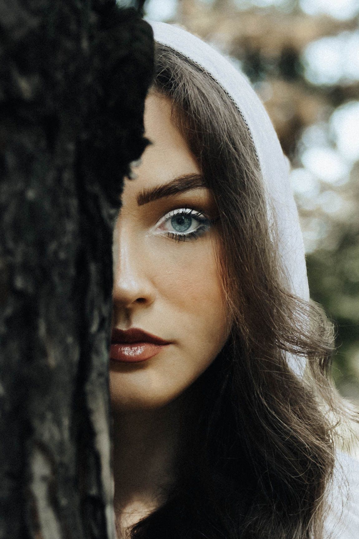 Close-up portrait with bright blue eyes and dark red lipstick against a blurred forest background.