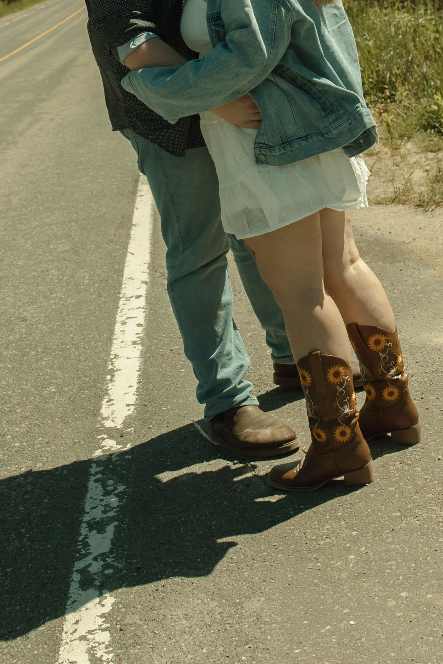Couple standing on road in cowboy boots, woman wearing sunflower boots and white skirt with denim jacket.