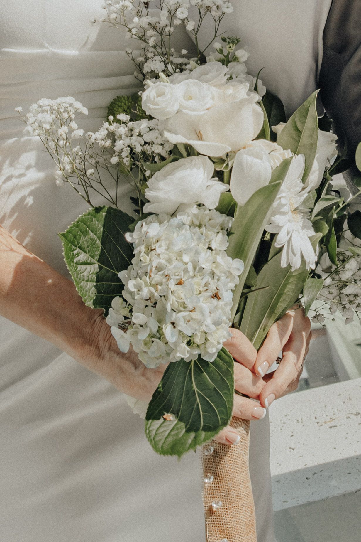 A bride and groom hold a elegant white wedding bouquet with roses, hydrangeas, and baby's breath.