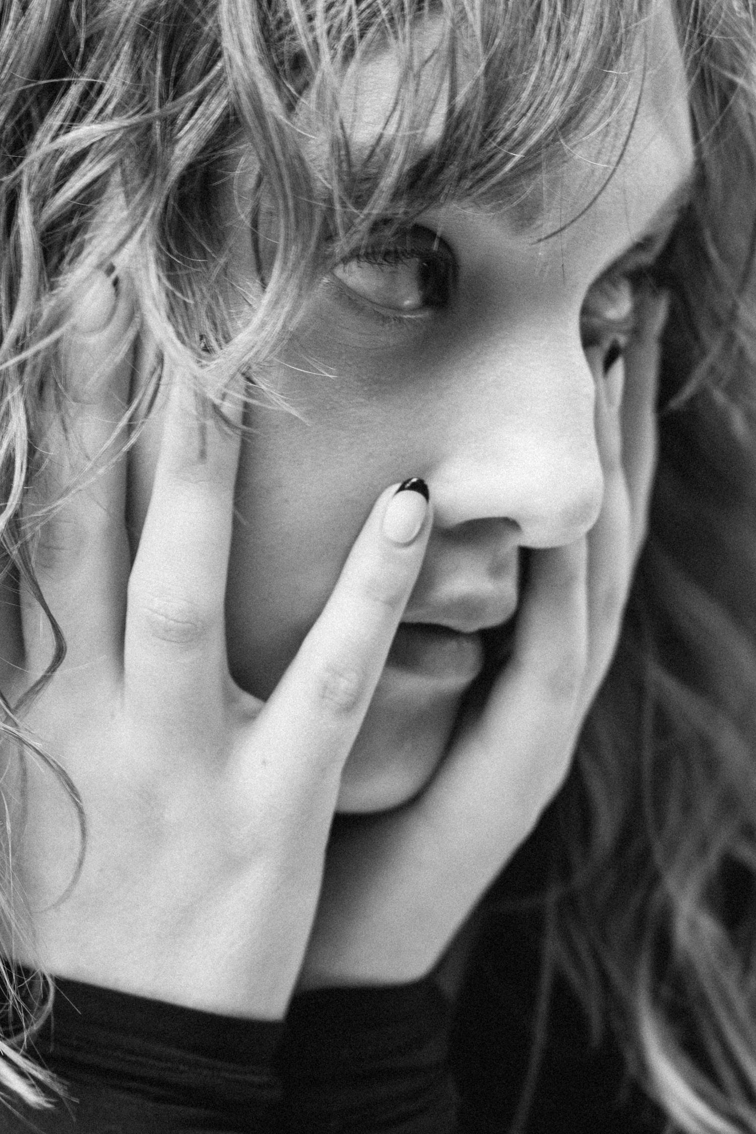 Black and white close-up portrait of a young woman with curly hair covering her face with her hand, dark nail polish.
