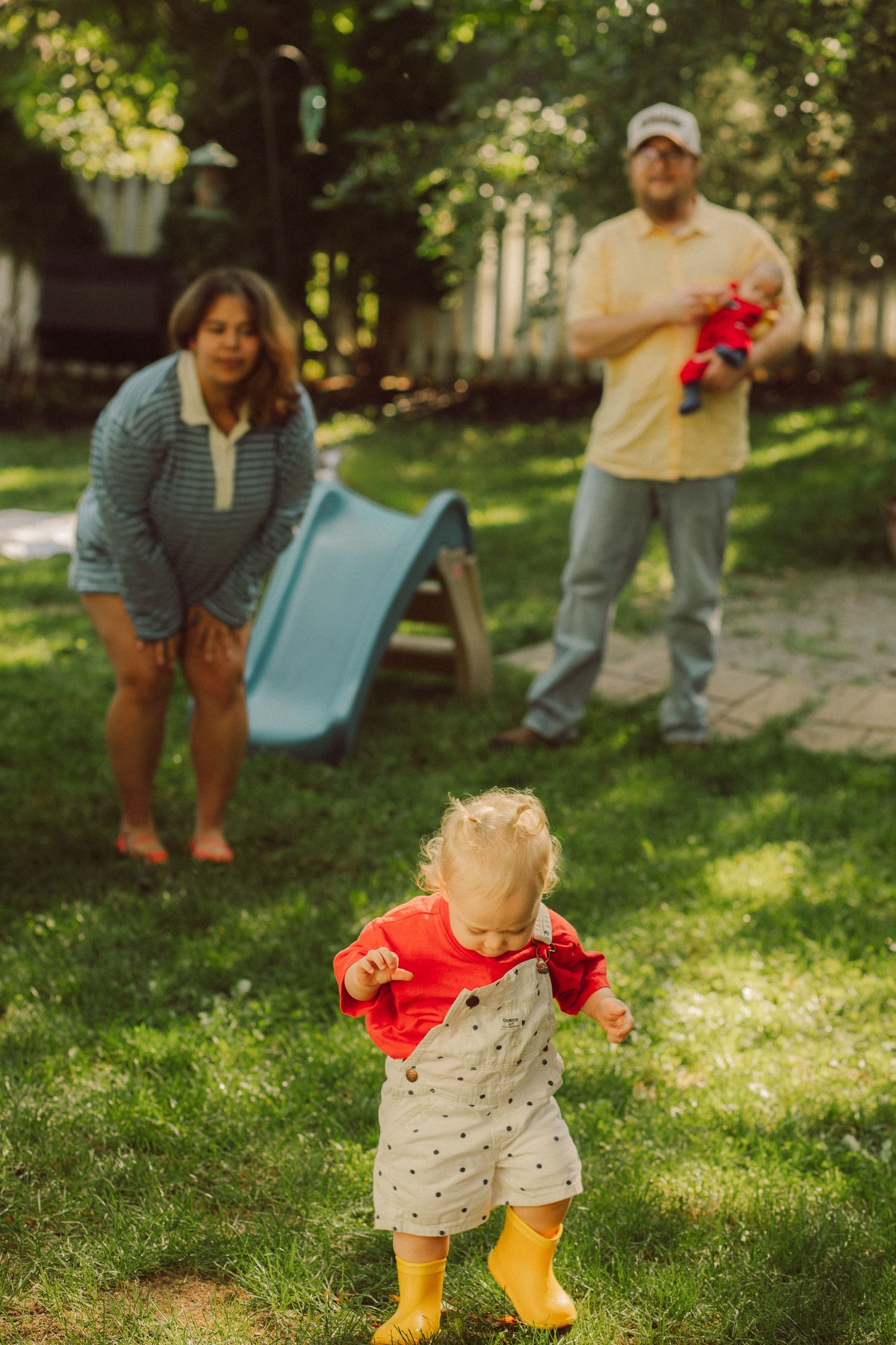 A toddler in a red jacket and yellow boots walks on grass while parents watch near a blue slide.