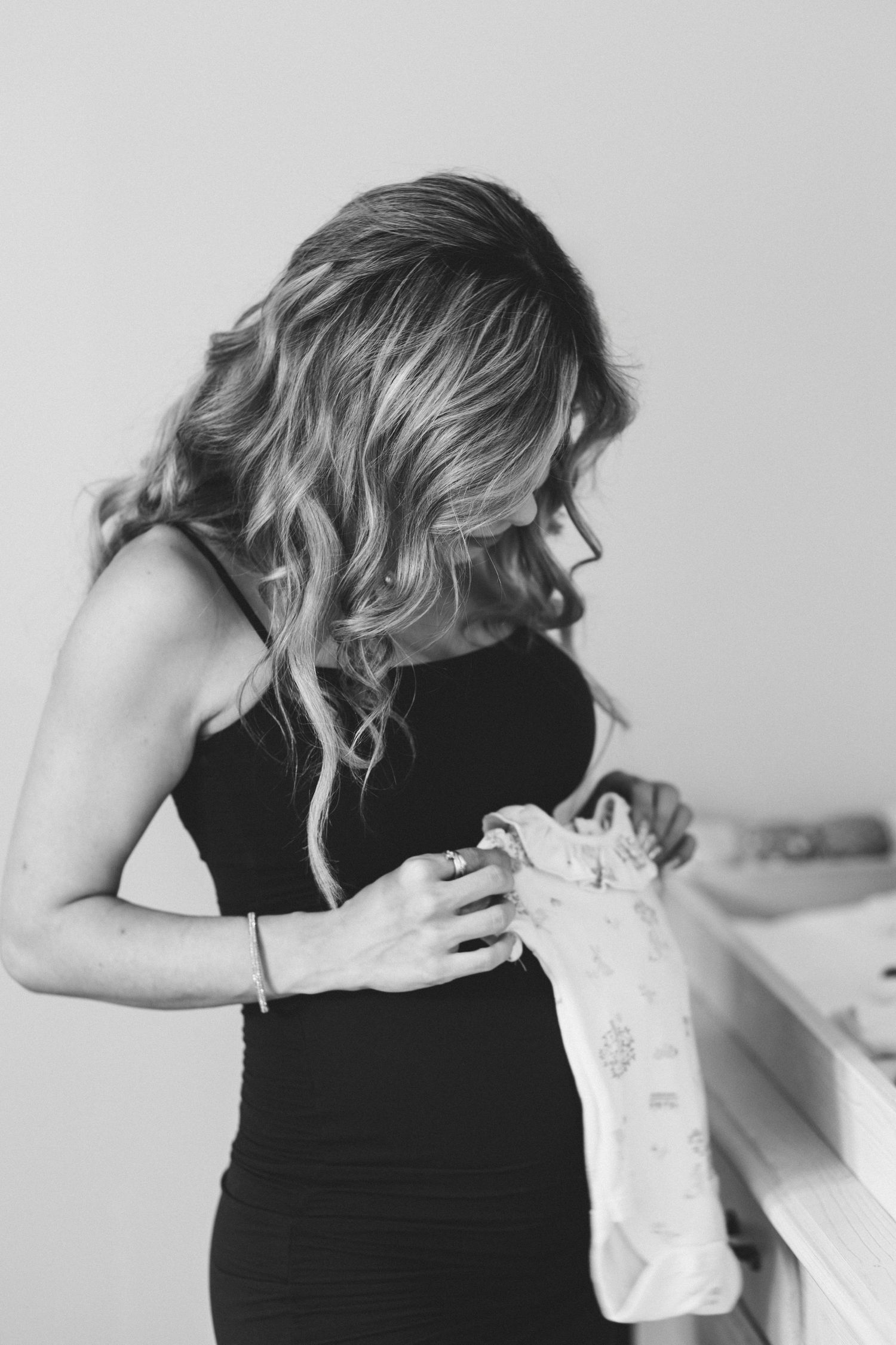 Pregnant woman in black dress holding baby clothes near a crib in black and white photo.