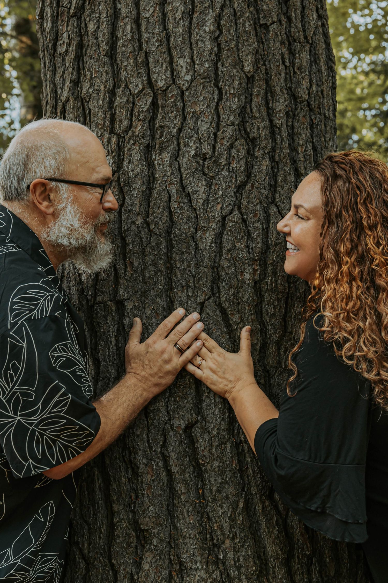 Two people playfully pose on opposite sides of a large tree trunk in the forest while wearing dark clothing.