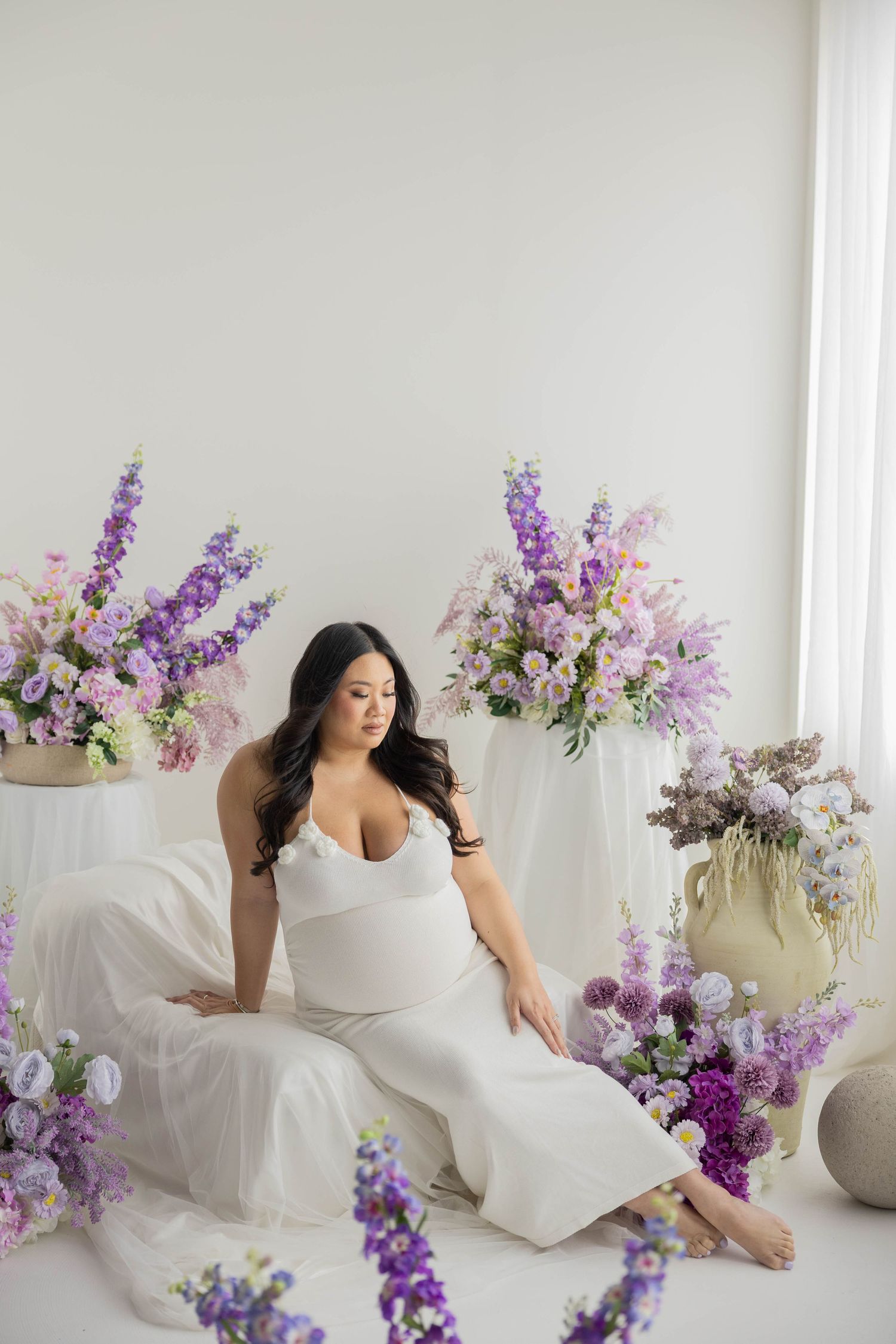 Artistic maternity portrait with elegant white draping and purple floral arrangements against light background.