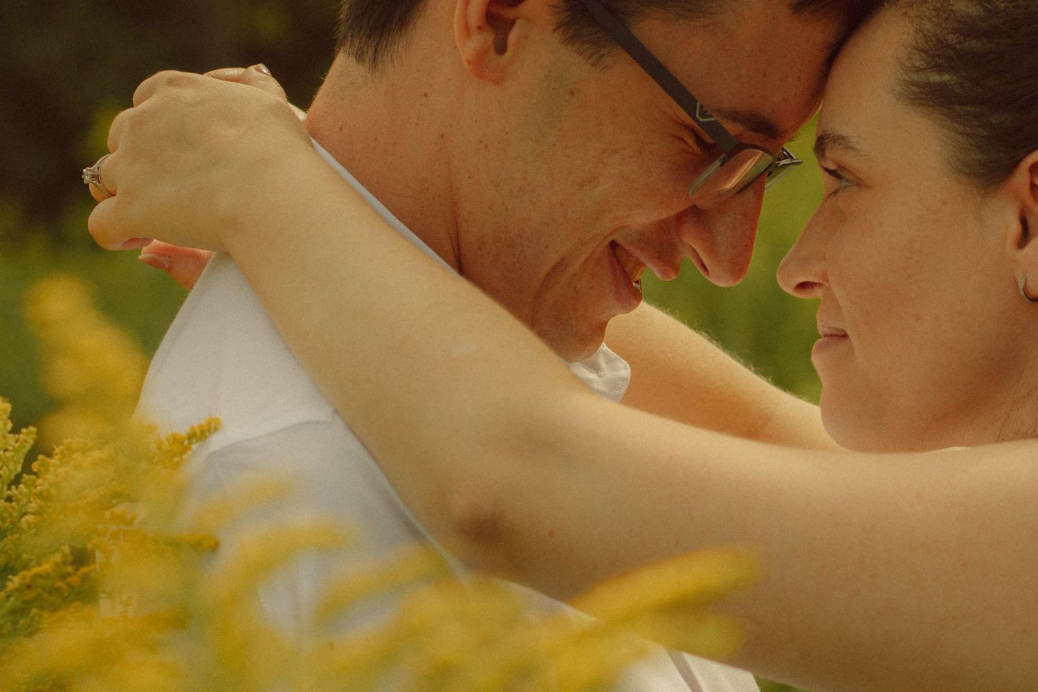 Romantic couple embracing outdoors among yellow wildflowers, man wearing glasses smiling at woman in warm natural light.