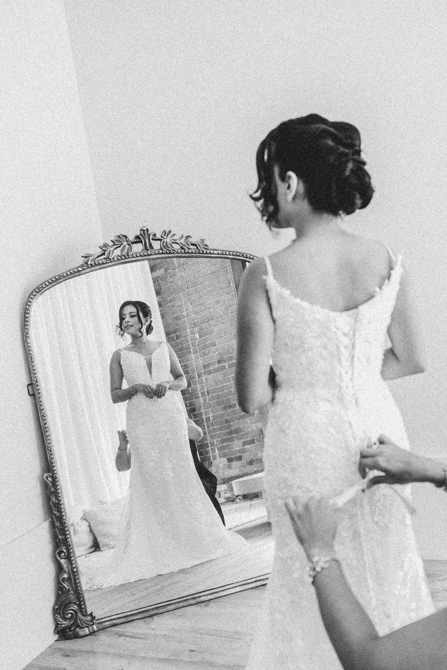 Elegant bride in a white lace wedding dress stands before an ornate vintage mirror showing her reflection.