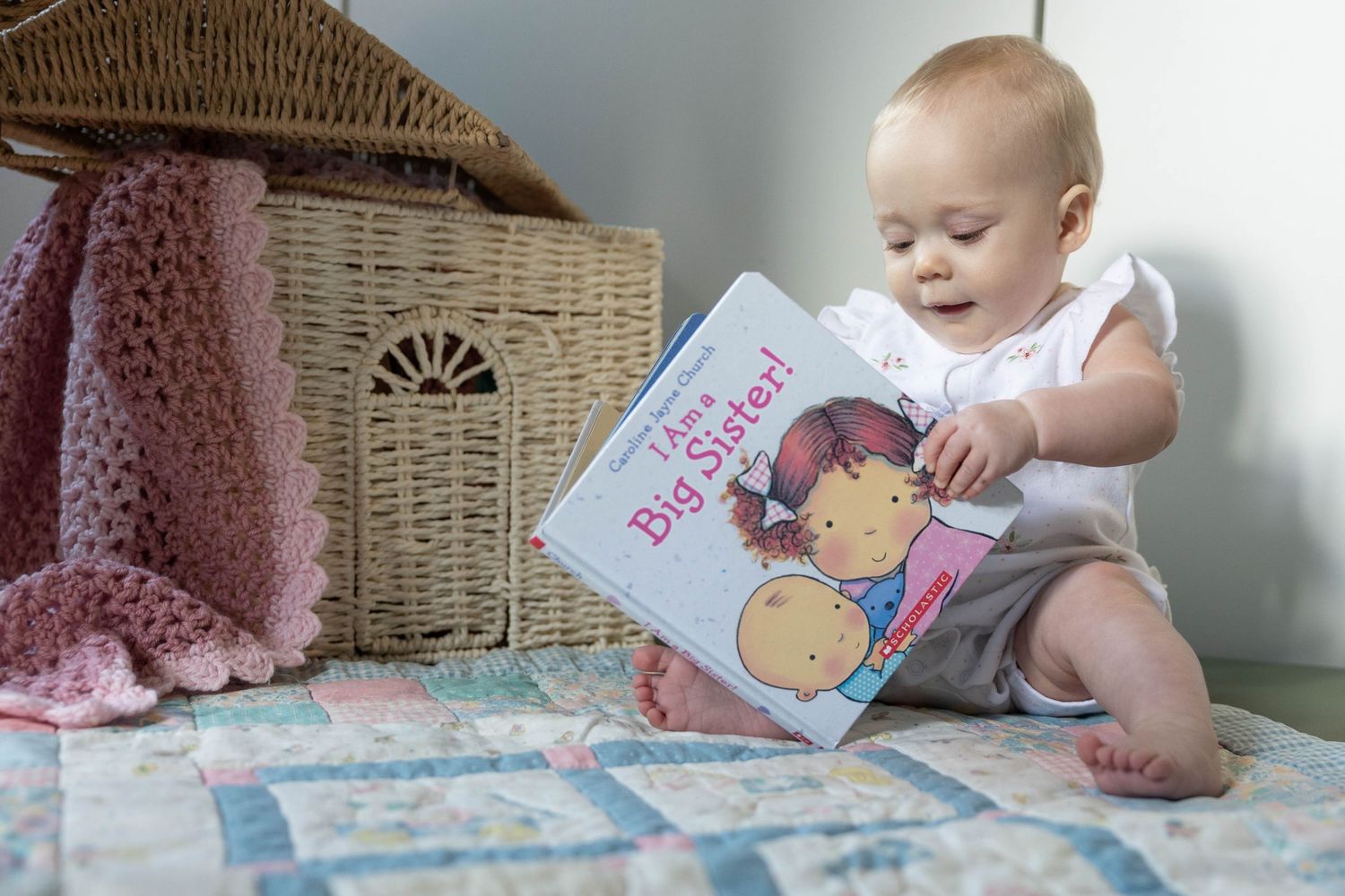 A baby in white clothing sits on a blanket looking at a children's board book.