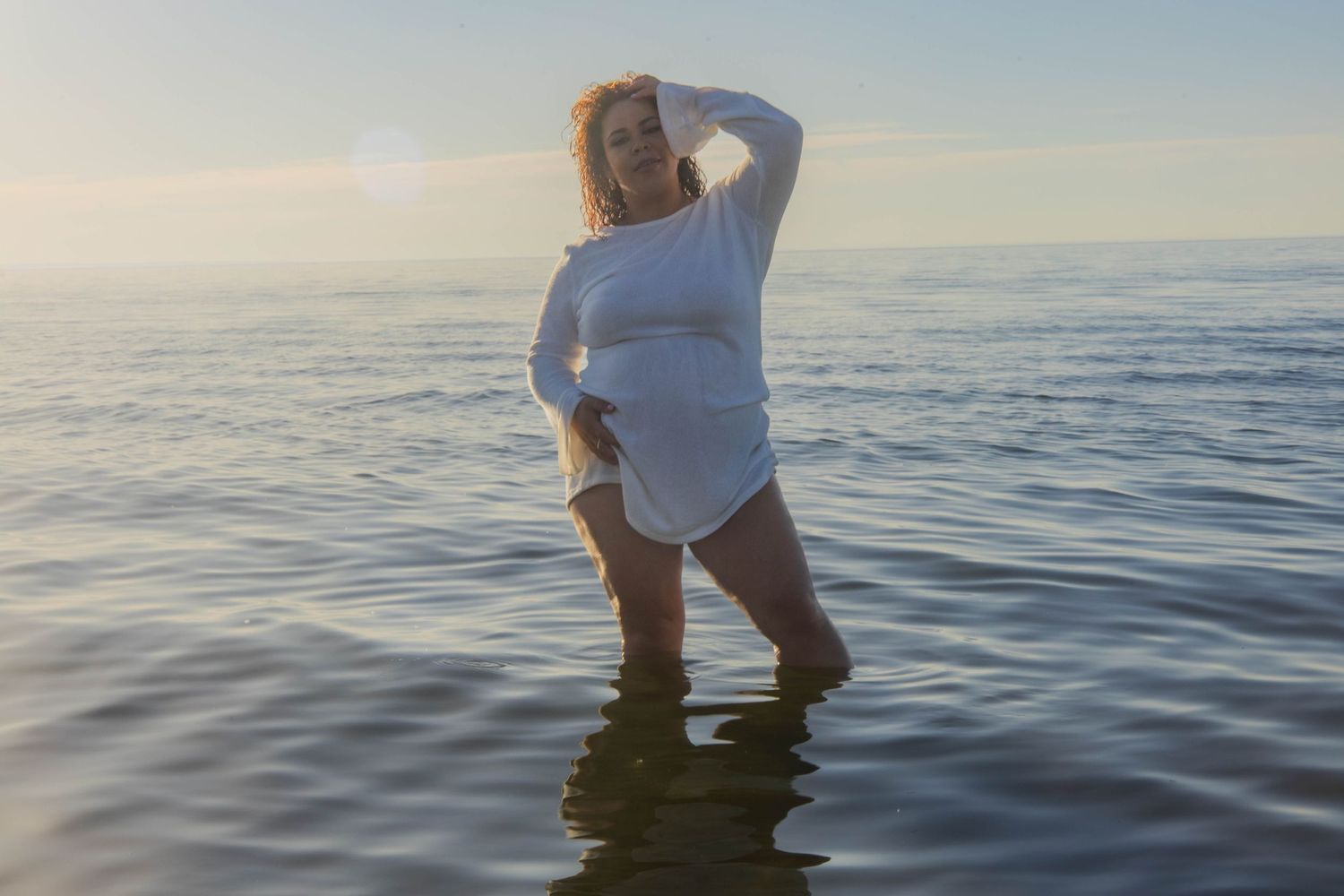 A figure wades in calm ocean waters at sunset creating rippling reflections.