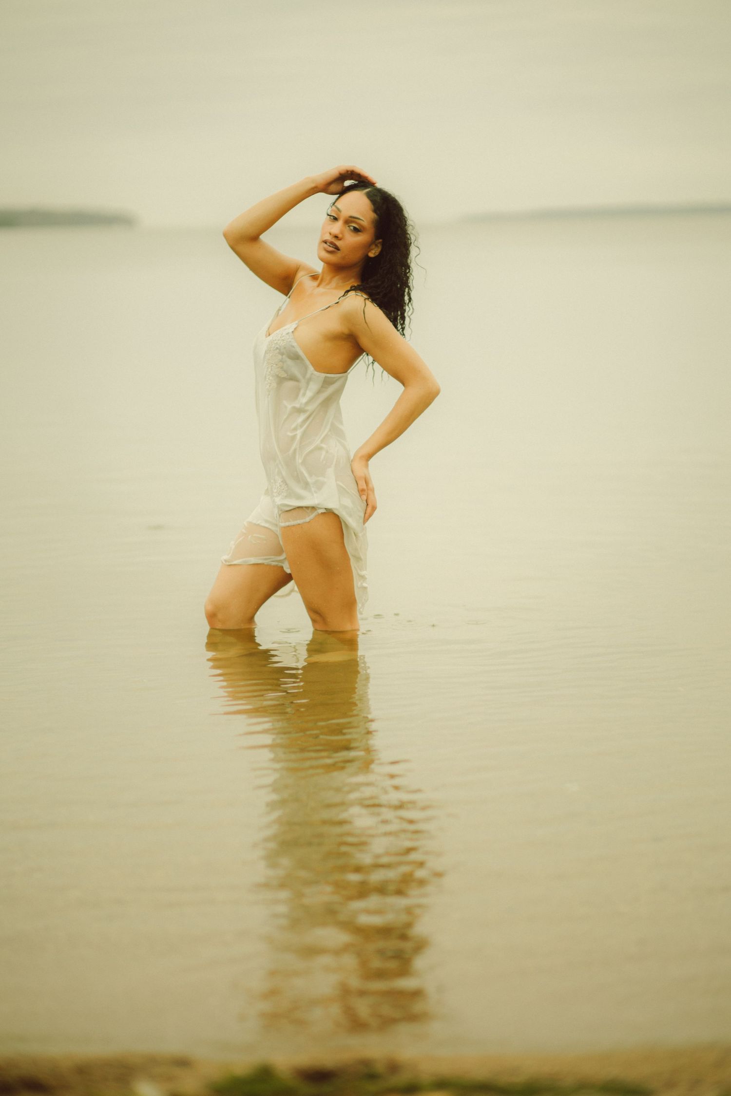 Woman in wet white dress standing knee-deep in calm water, posing with hand in hair on overcast day.