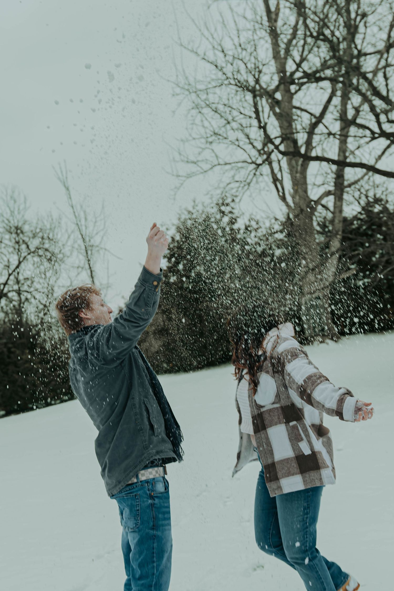 Two people playfully throw snow at each other in a winter landscape wearing jeans and jackets.