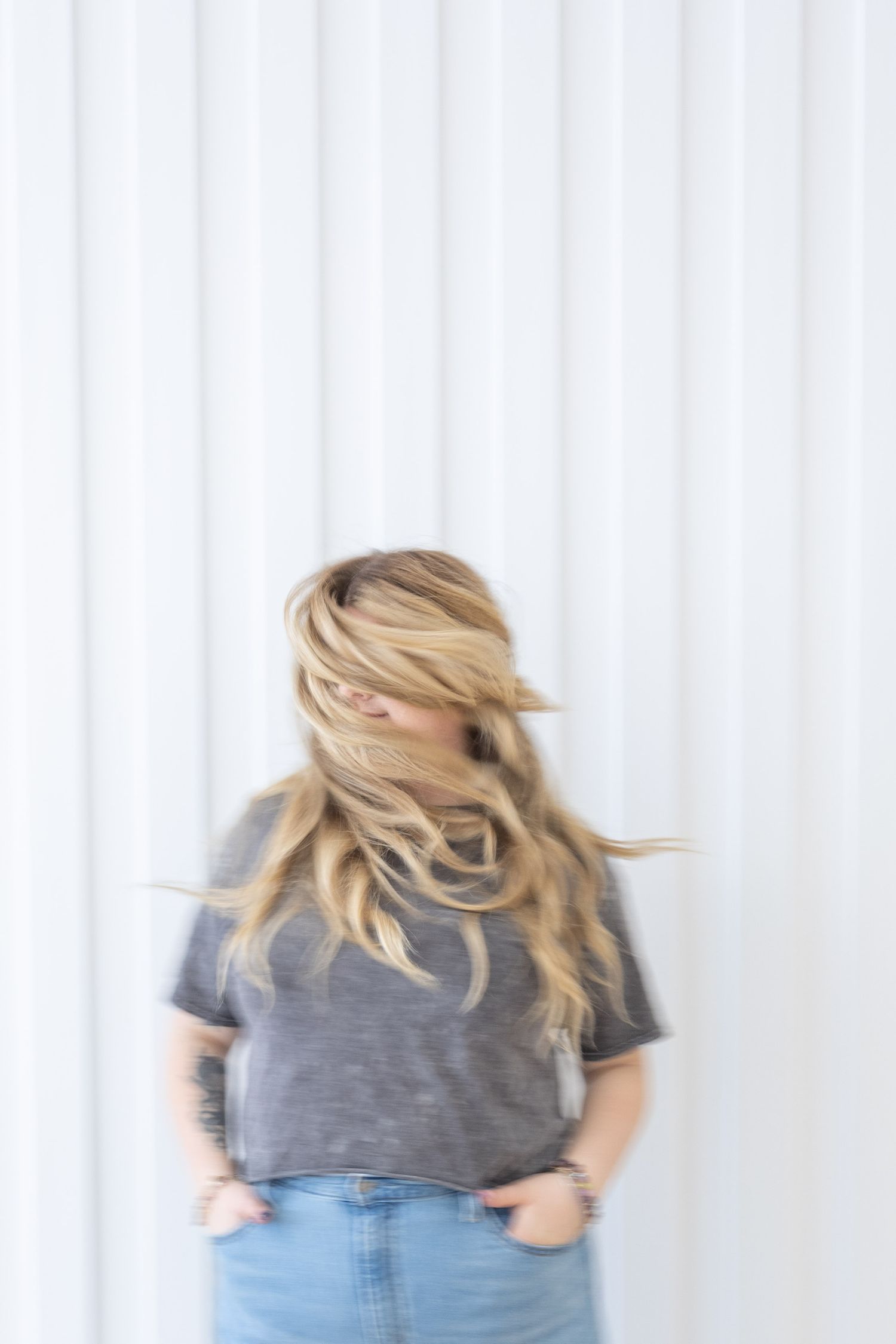 Blonde woman in gray t-shirt and jeans with hair blowing across her face against a white background.