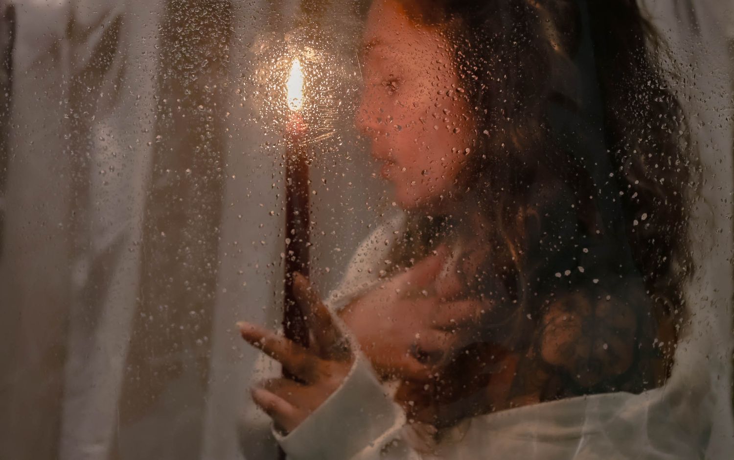Contemplative portrait through rain-covered window with soft ambient light.