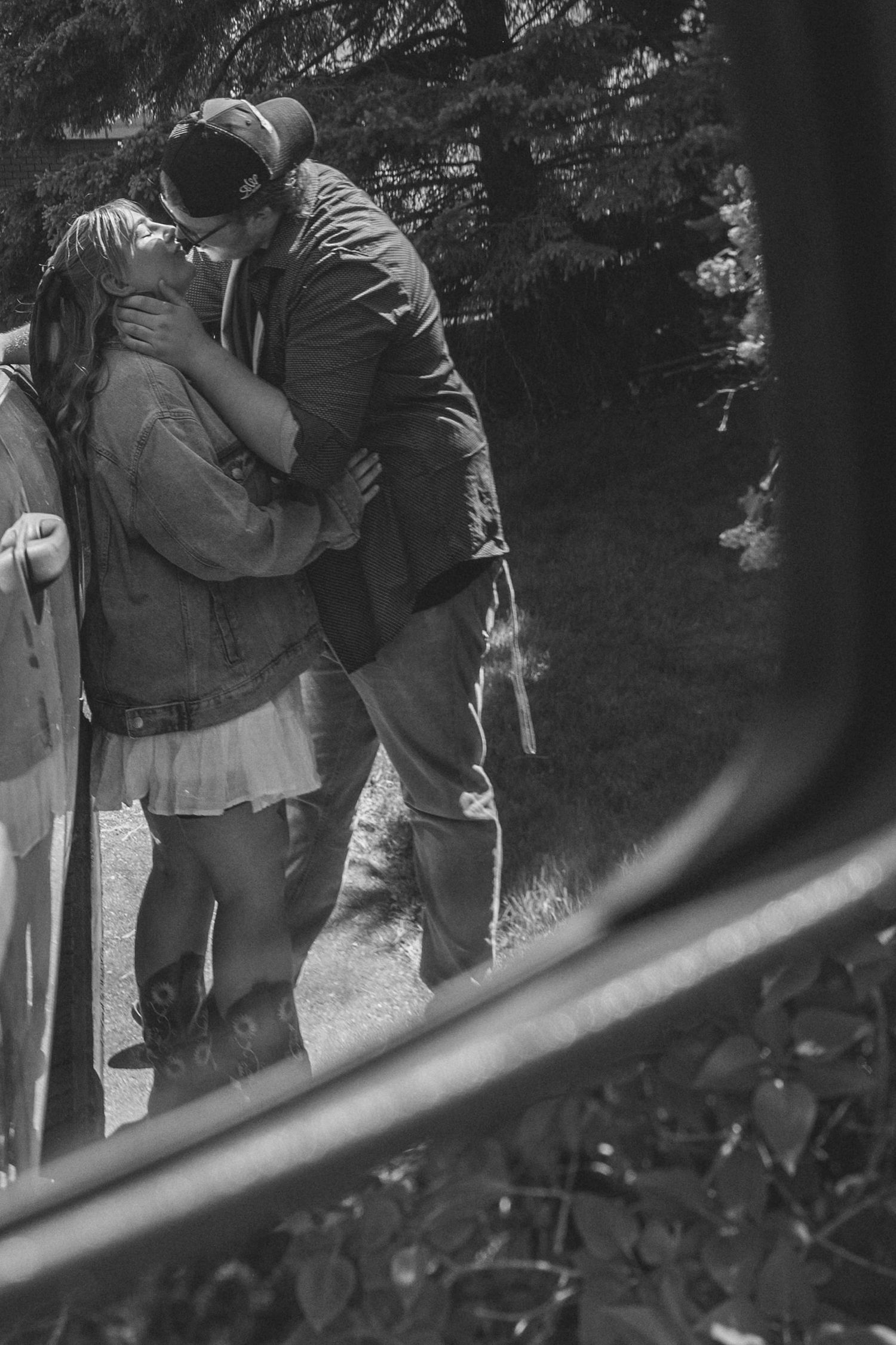 Black and white photo of a couple embracing outdoors in a natural wooded setting with trees in the background.