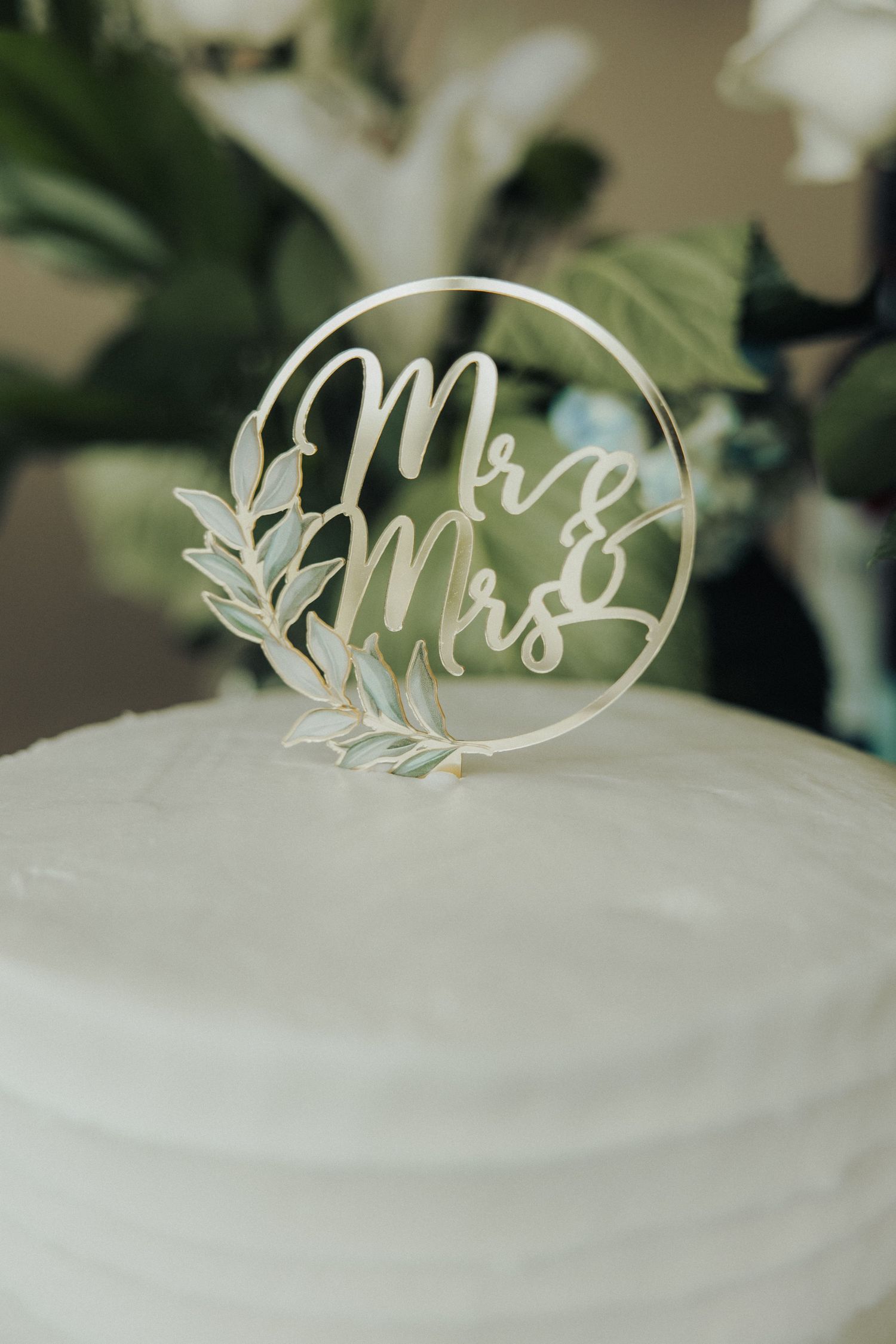 Elegant white wedding cake topped with a circular Mr & Mrs acrylic cake topper with leaf accents.