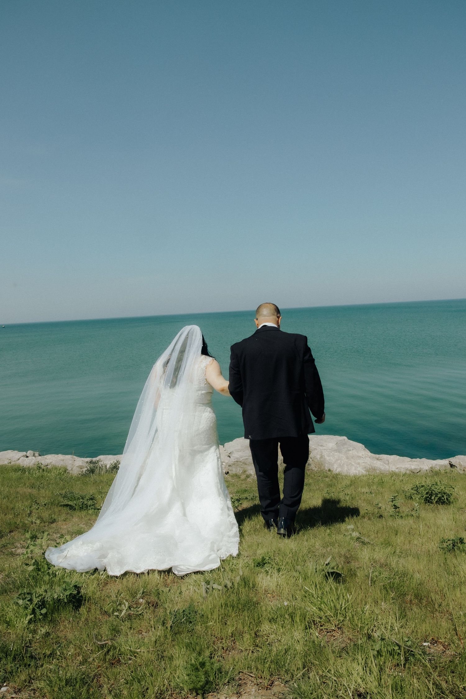 Bride and groom standing at cliff edge overlooking turquoise ocean, bride in white gown with veil, groom in black suit.
