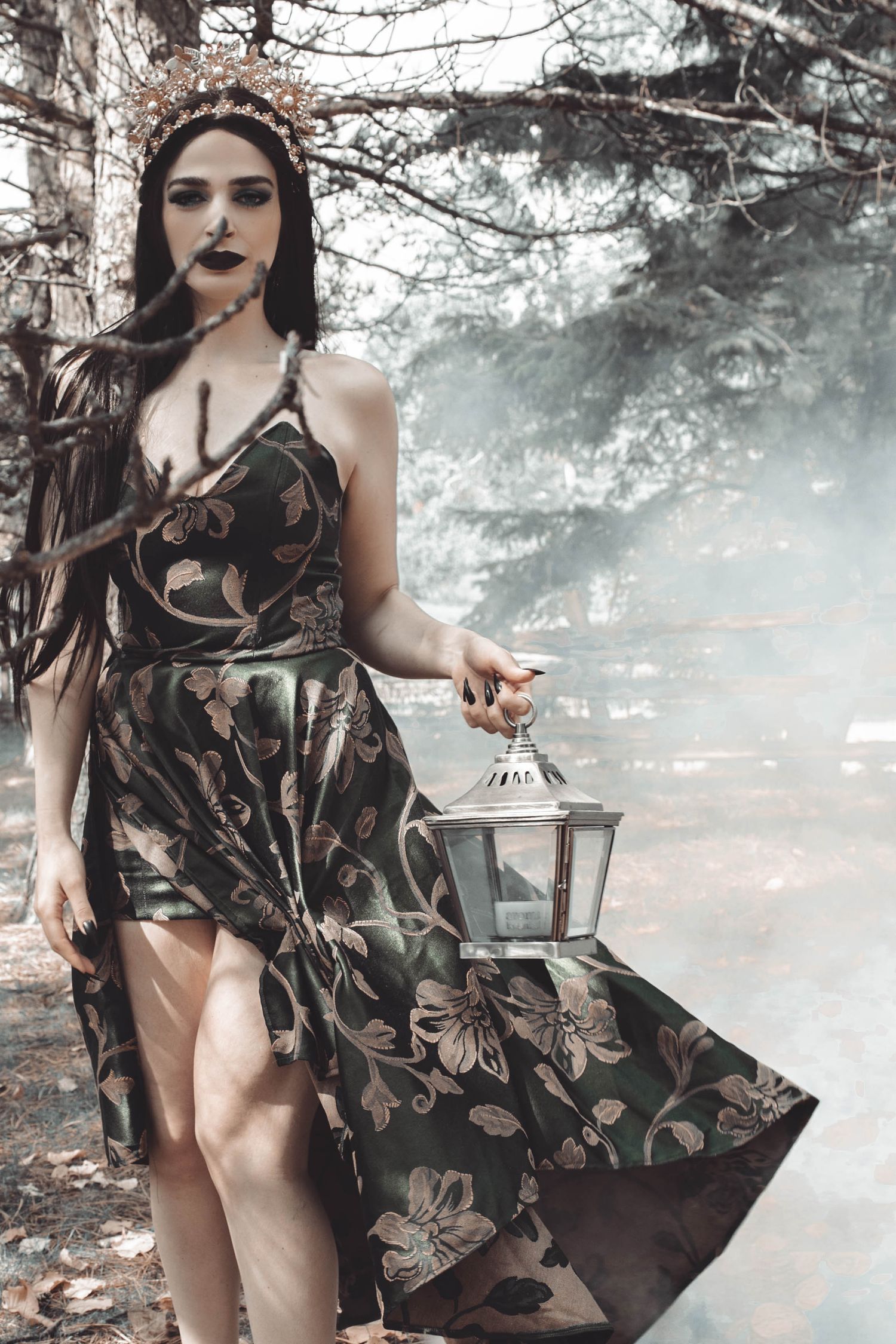 A dark floral dress with a high slit flows dramatically in a misty forest setting while holding an antique lantern.