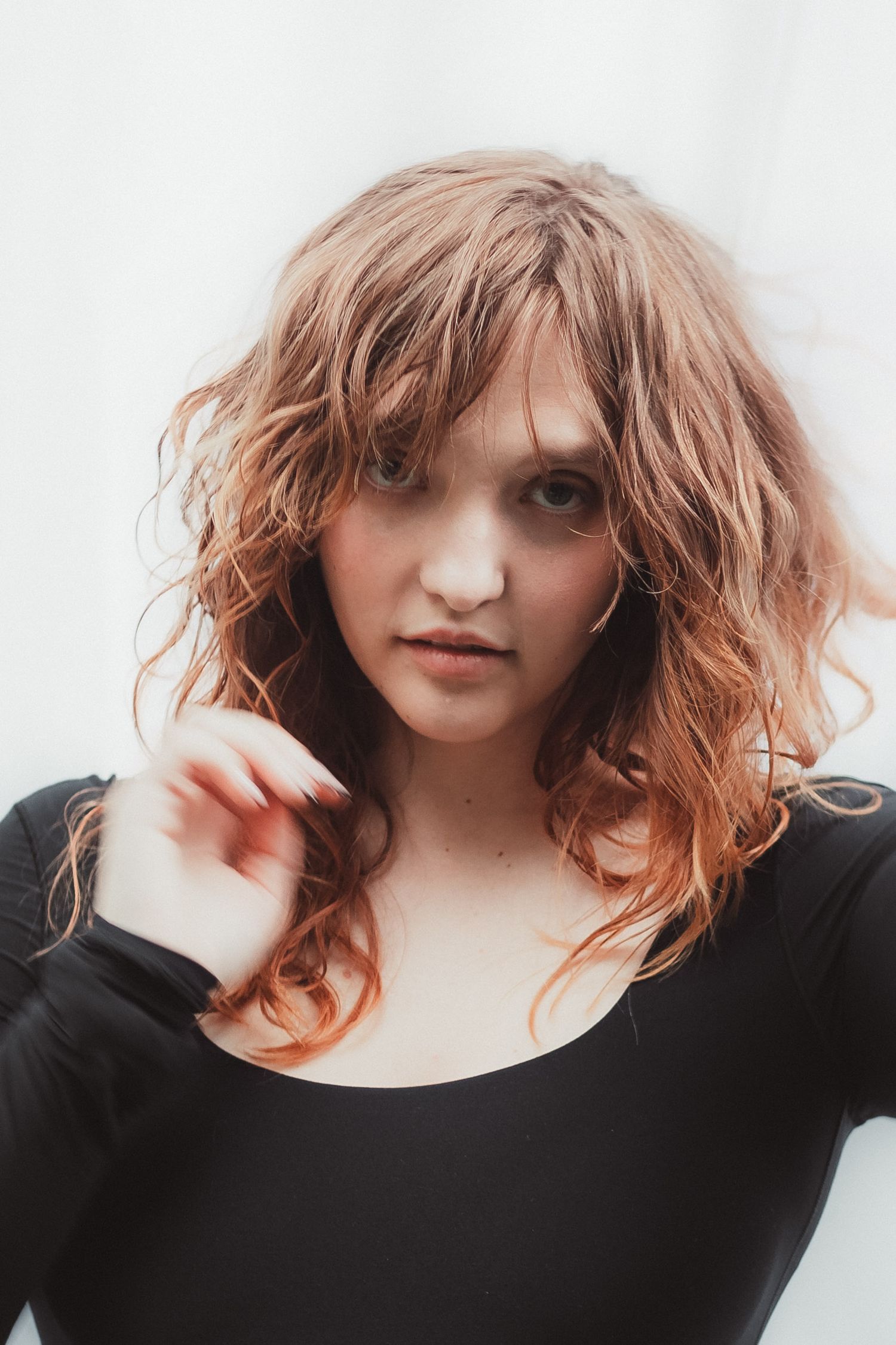 Young woman with curly auburn hair and bangs wearing a black long-sleeve top, posing against a white background.