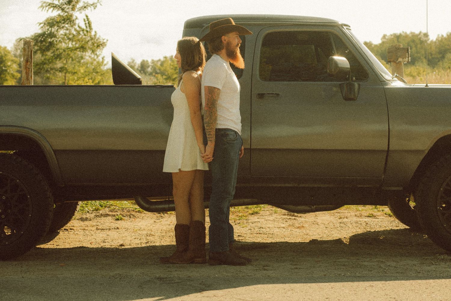Couple standing back-to-back by a classic pickup truck outdoors on a sunny day.