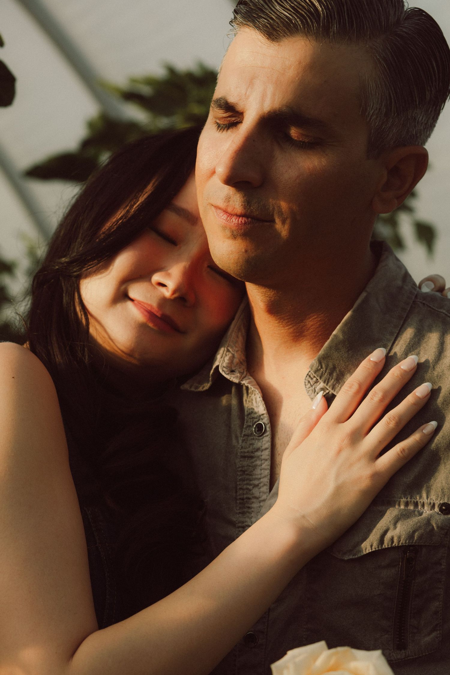 A couple embraces tenderly in warm golden light, woman resting her head on man's chest with eyes closed.