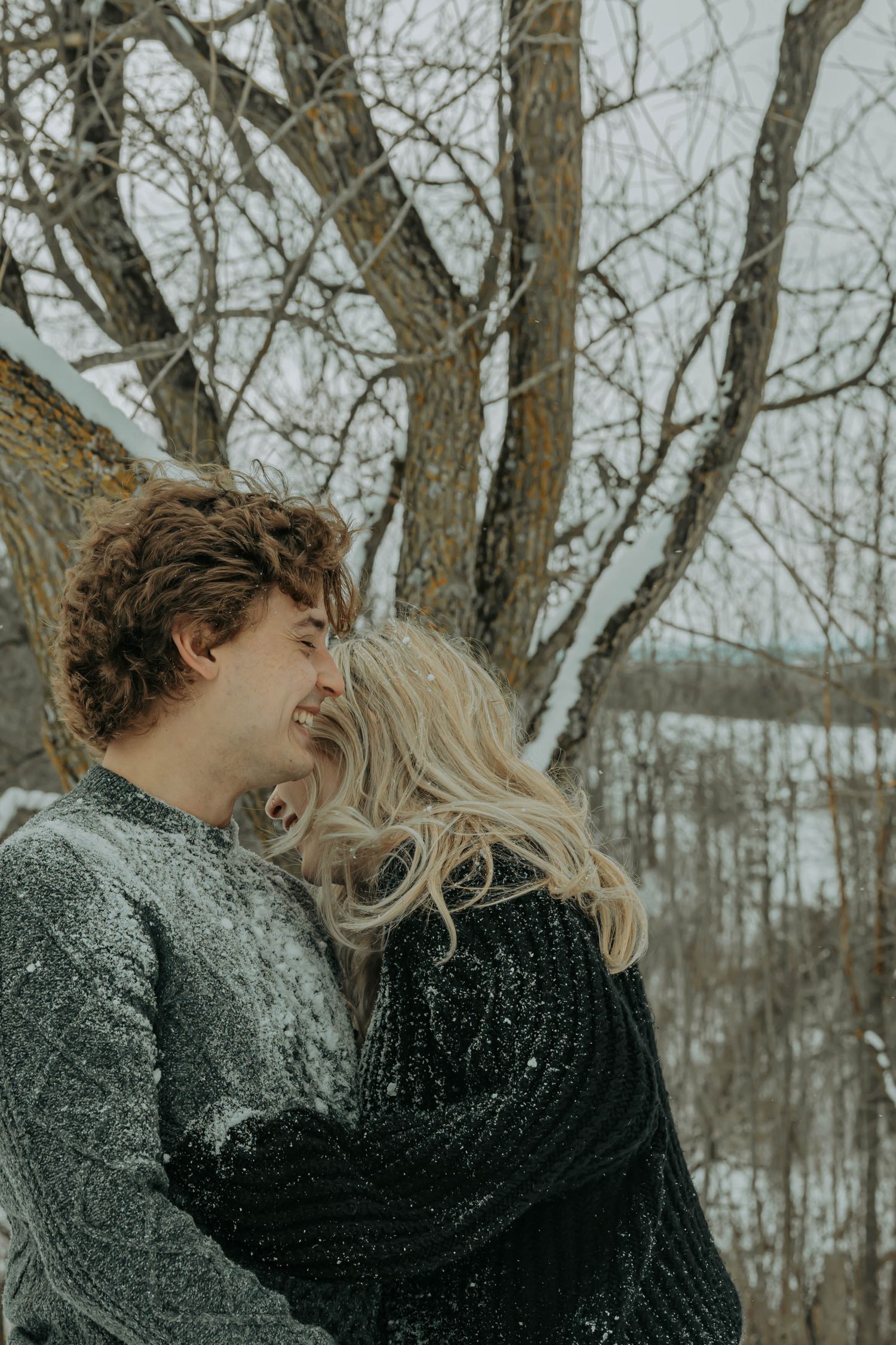 A couple embraces and laughs together in a winter forest setting with bare trees in the background.