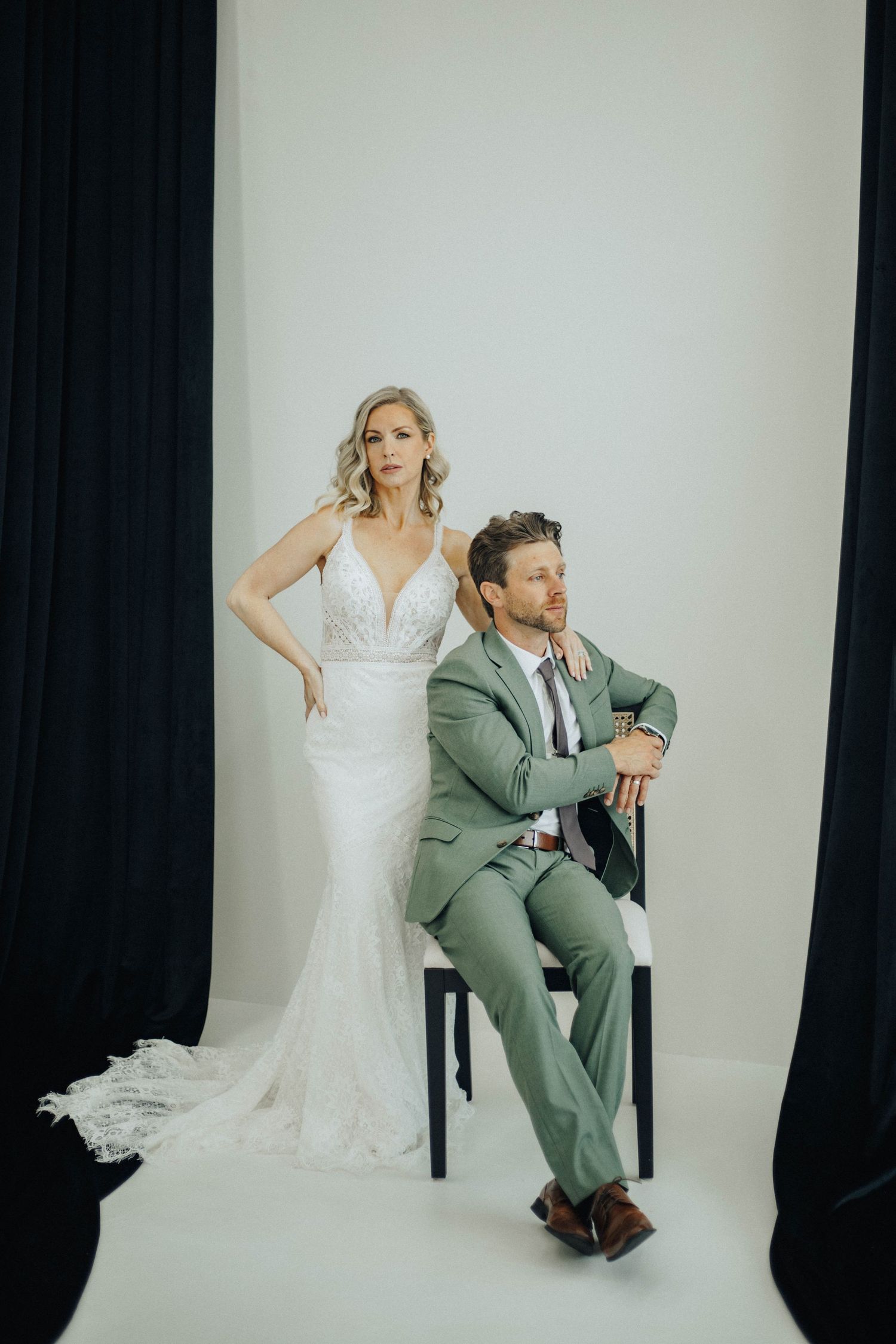 A wedding couple poses playfully in a studio setting with a bride in white dress and groom in sage green suit.