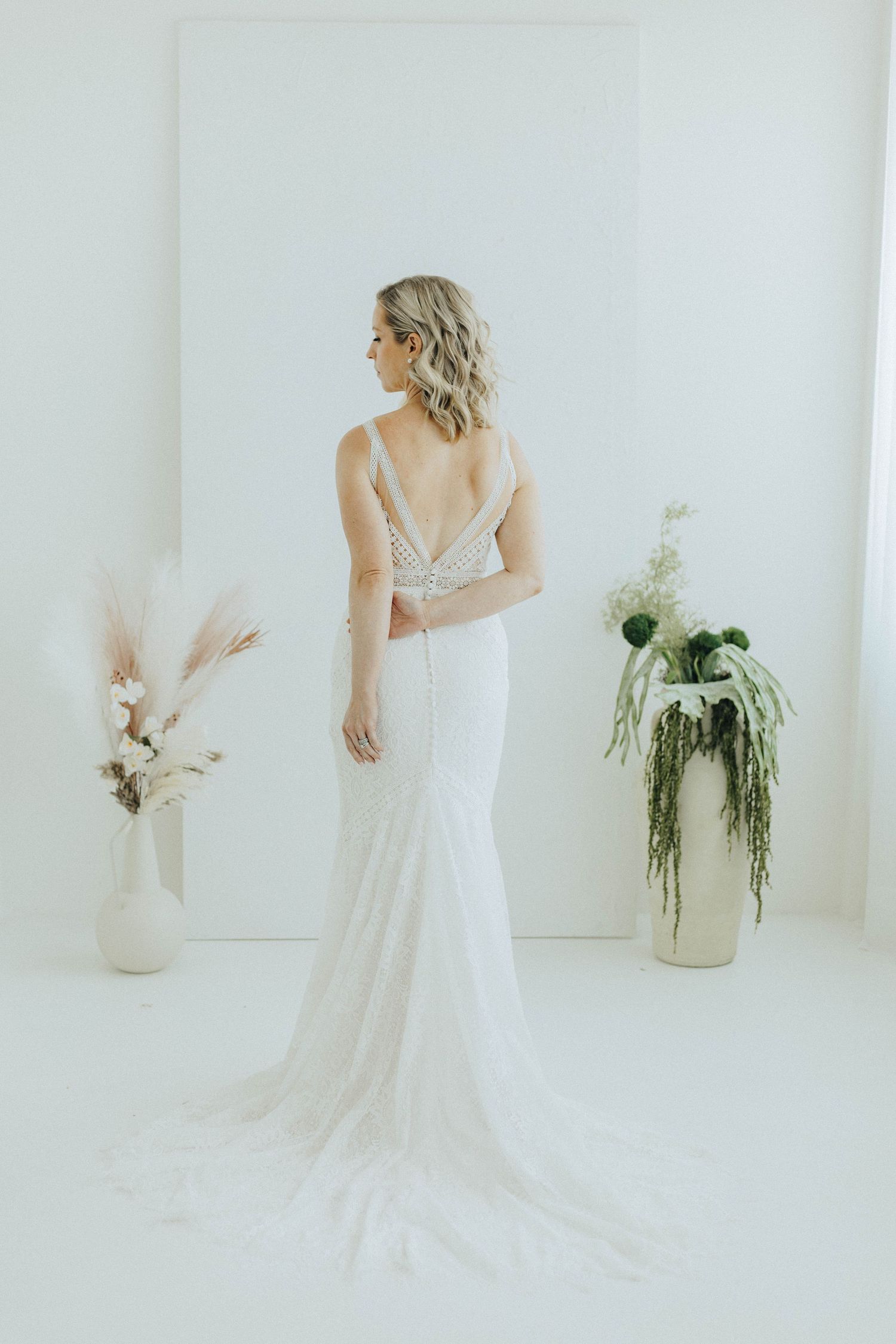 Elegant white wedding dress with plunging back and train shown in minimal studio setting with dried floral arrangements.