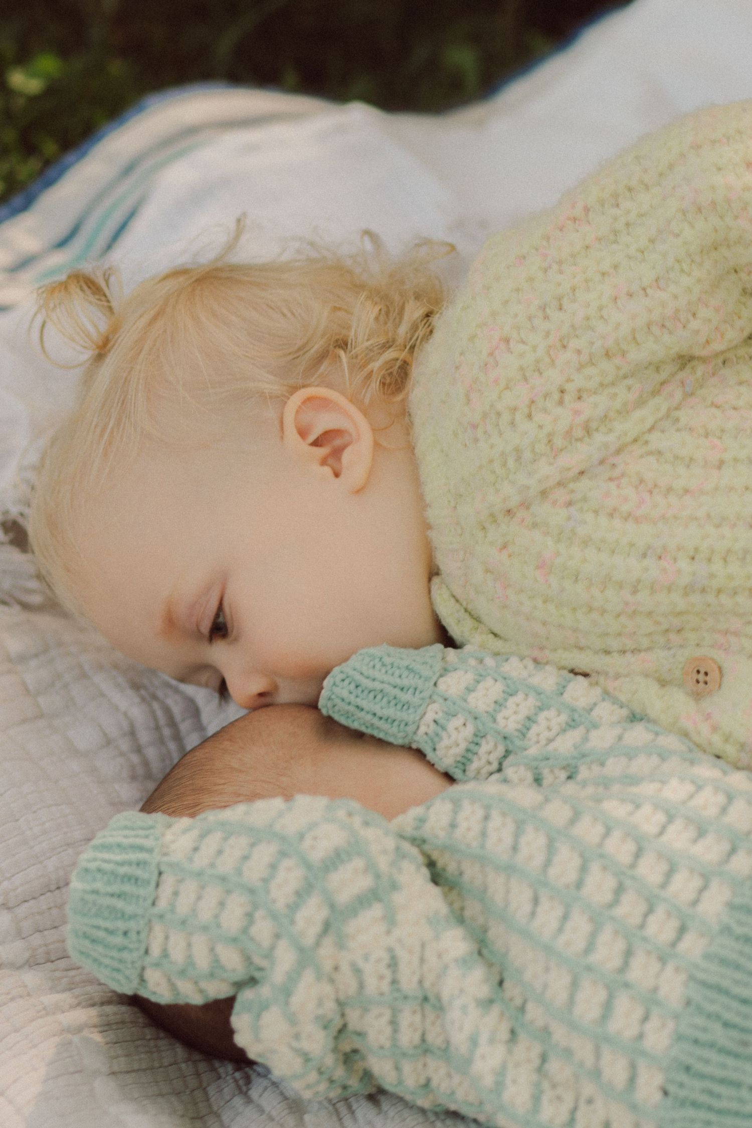 Blonde toddler in mint knit sweater breastfeeding outdoors on a soft blanket in natural light.