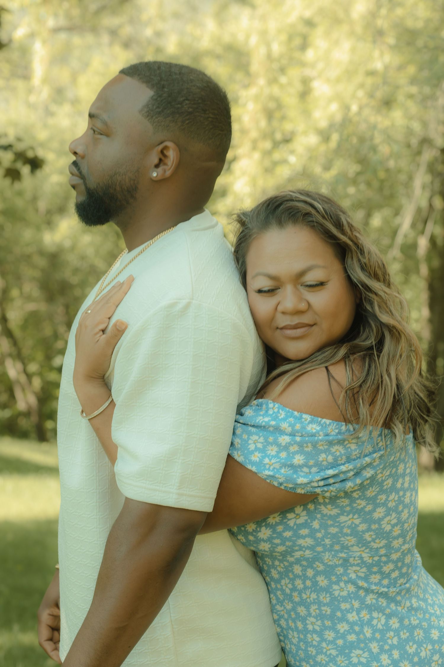 A woman in a blue floral dress embraces a man in a white shirt from behind in a lush green park.