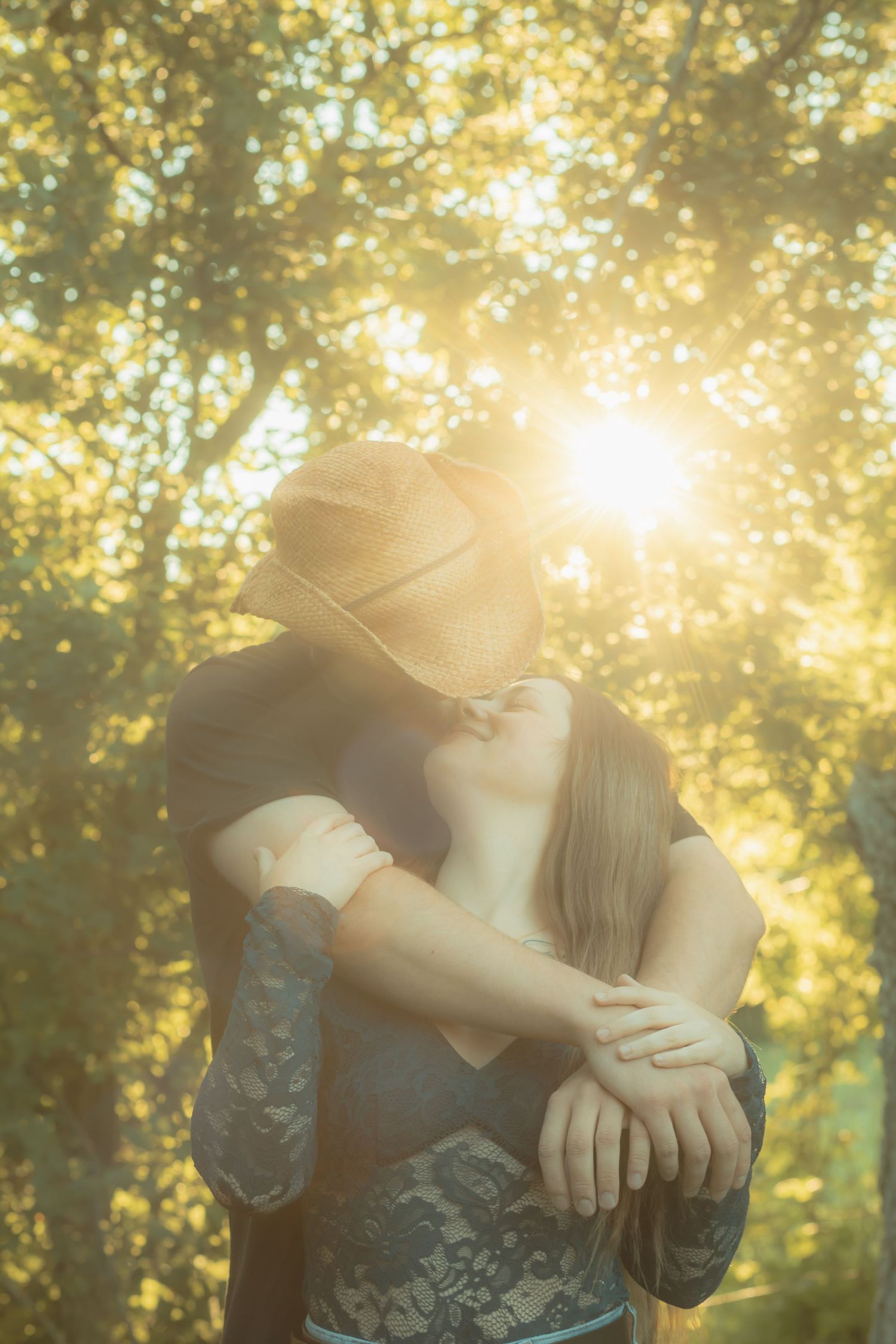 Couple embracing and kissing in golden sunlight filtering through lush green trees outdoors.