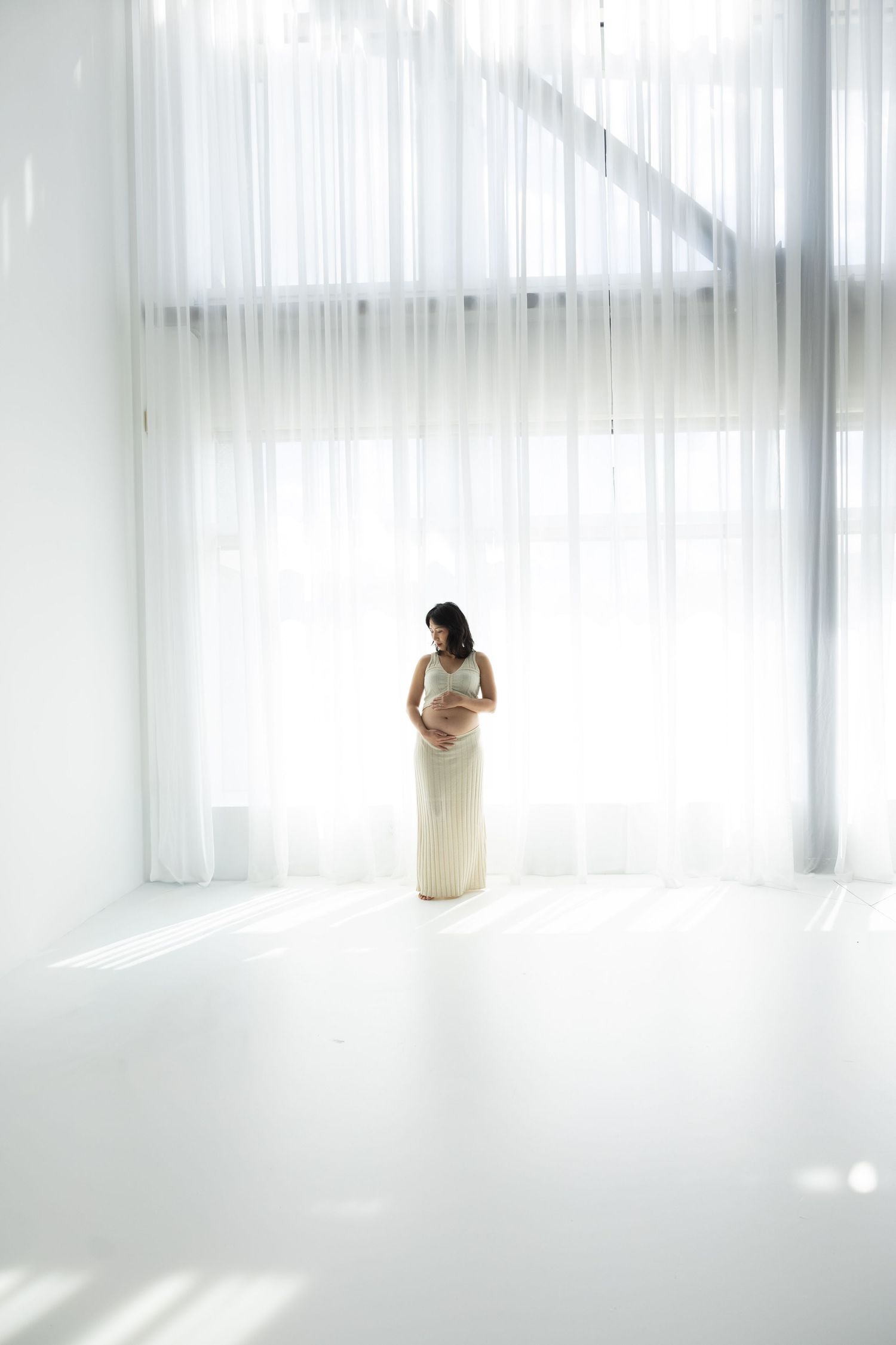 Pregnant woman in white dress cradling her bump, standing in a bright minimalist room with sheer curtains.