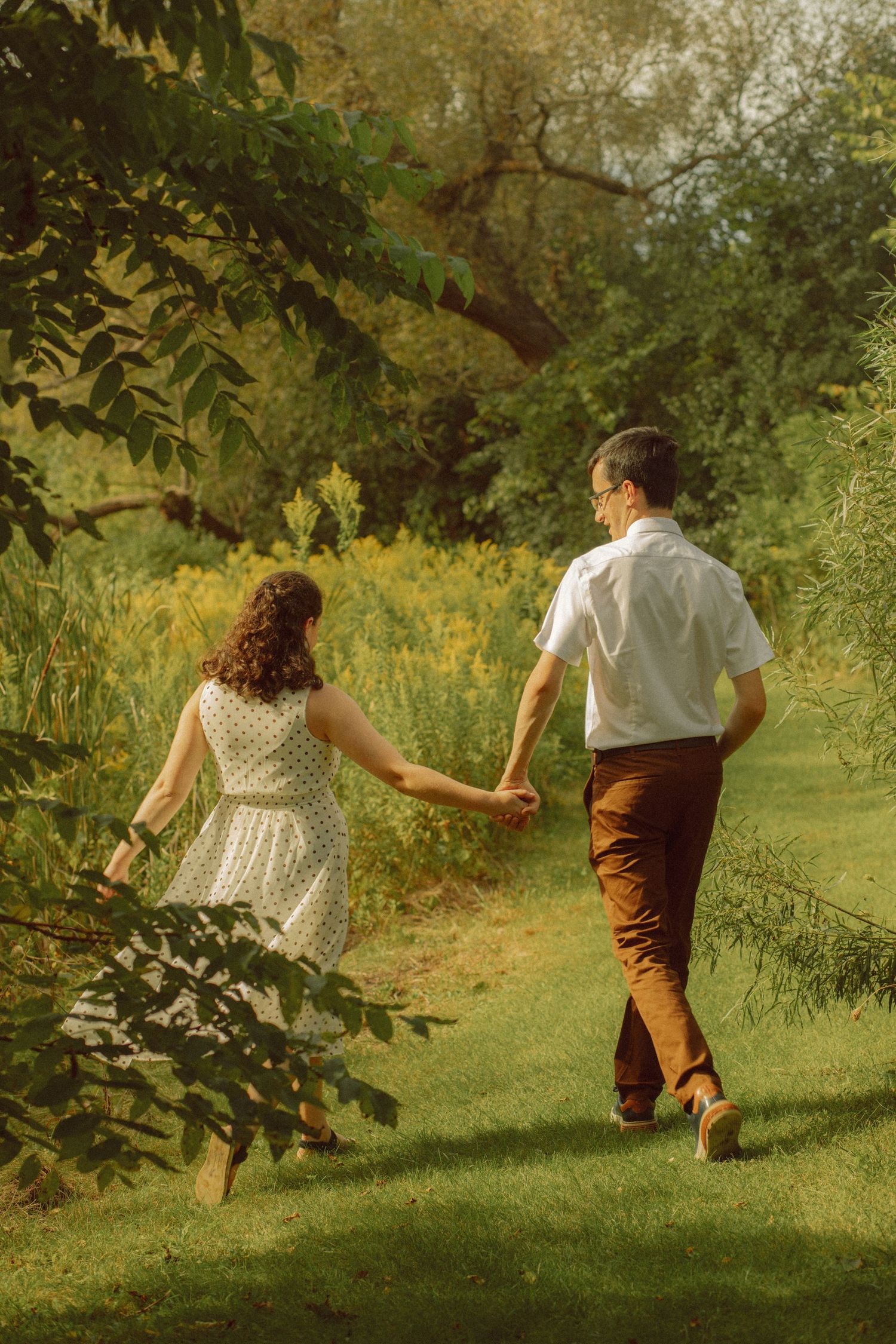 Couple holding hands walking through a lush green park path in golden sunlight.