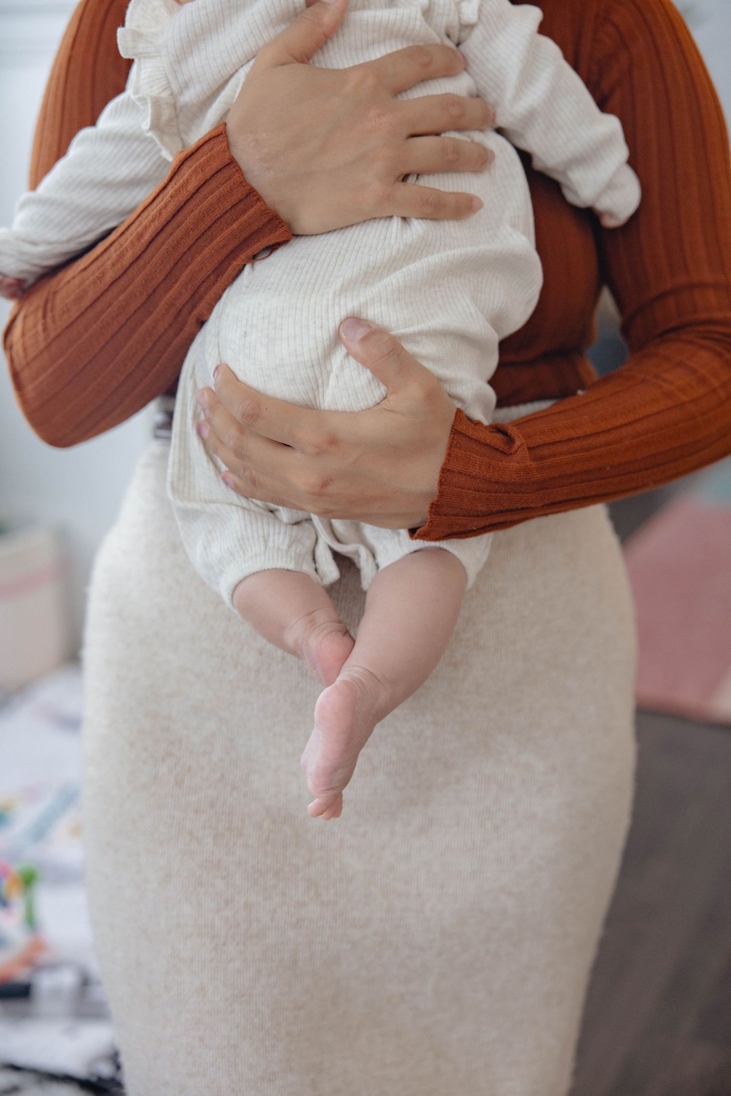 Baby in white outfit rests on a beige blanket while being held by someone wearing a rust-colored sweater.