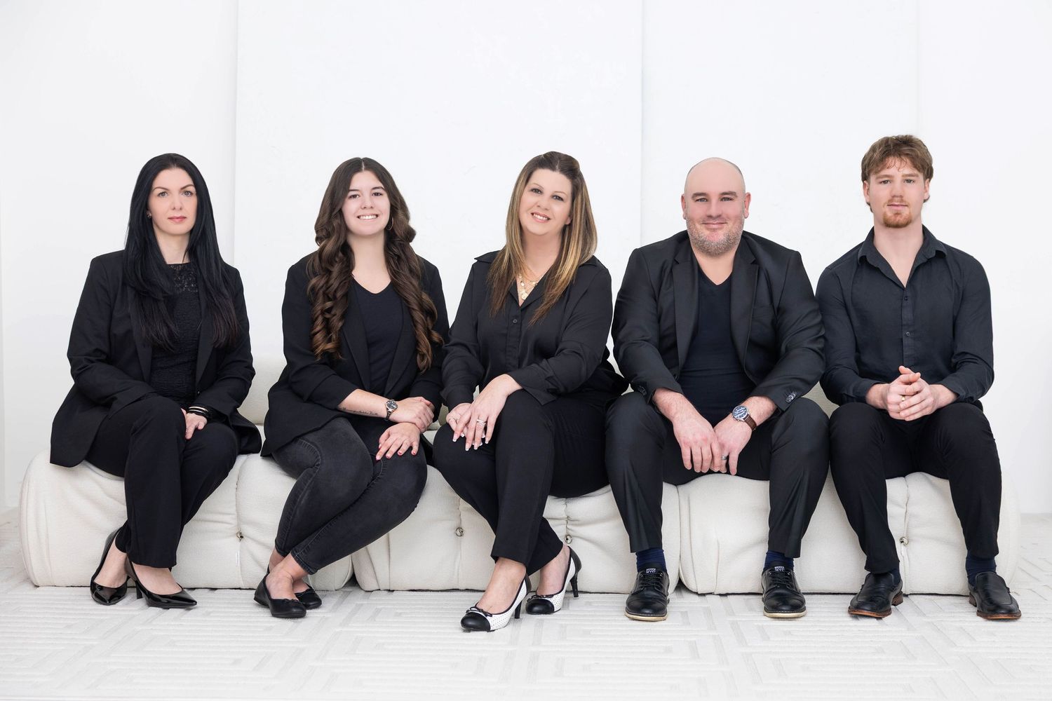 Group of people wearing black clothing sit casually in a row against a white background.