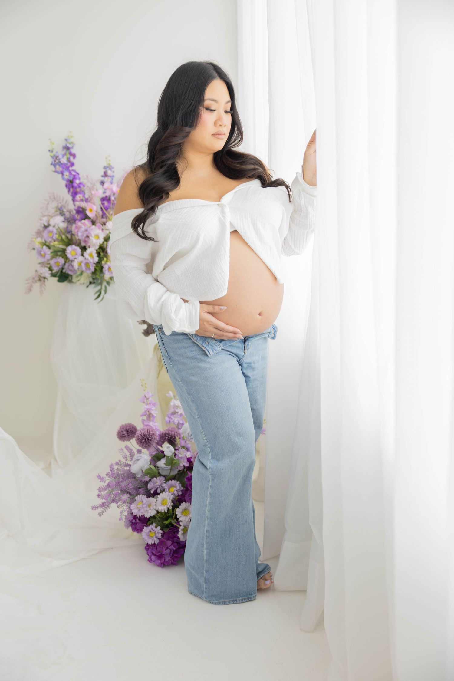 Ethereal maternity photoshoot with white fabric and purple floral arrangements in a bright studio setting.