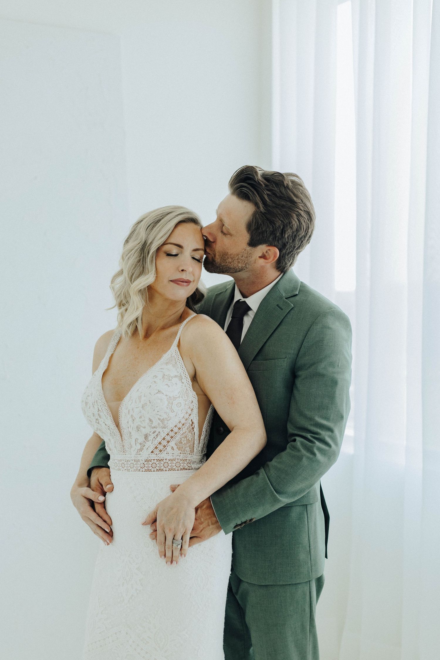Romantic wedding photo with couple in elegant attire embracing against white backdrop in soft natural light.