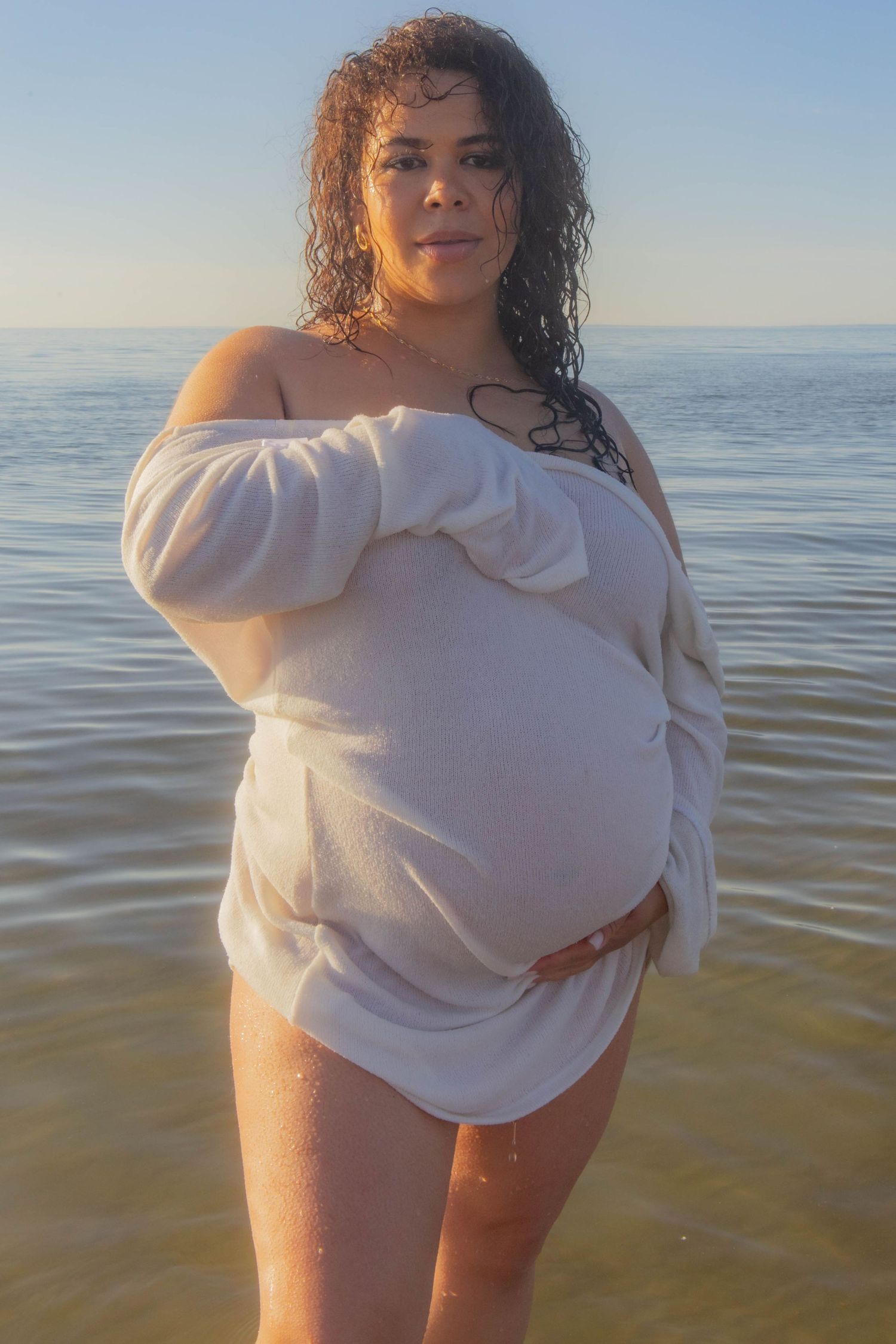 Person in white sweater poses in shallow ocean water during daytime.