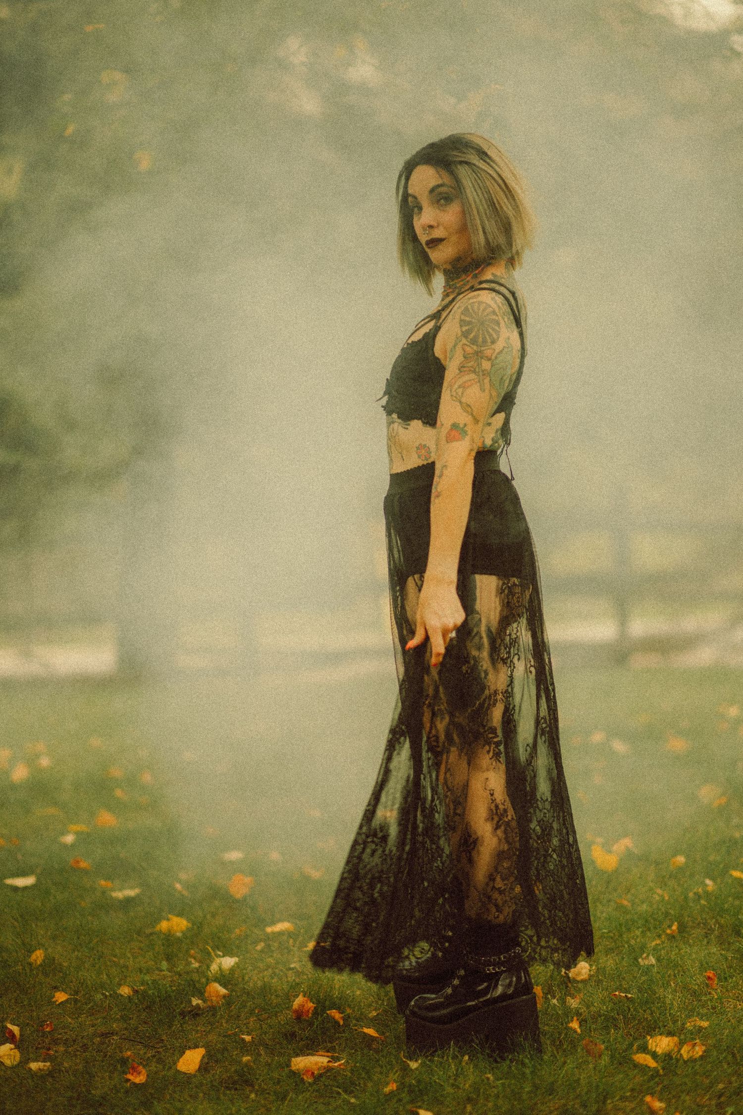 Tattooed woman in black lace skirt and crop top poses in misty autumn park with fallen leaves.