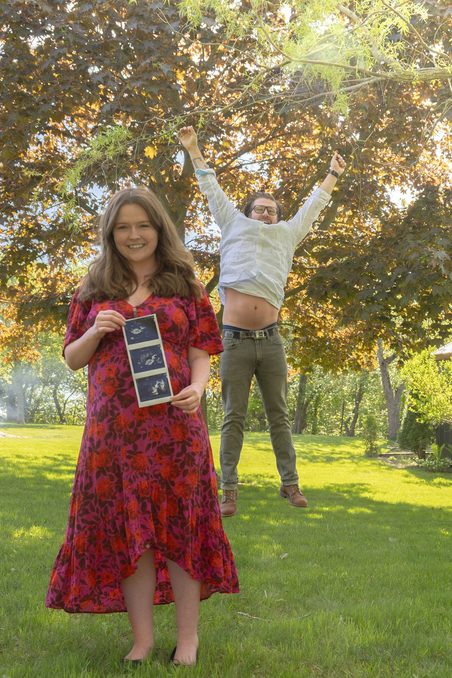 Pregnant woman in red floral dress holds ultrasound photos while excited partner celebrates behind her outdoors.