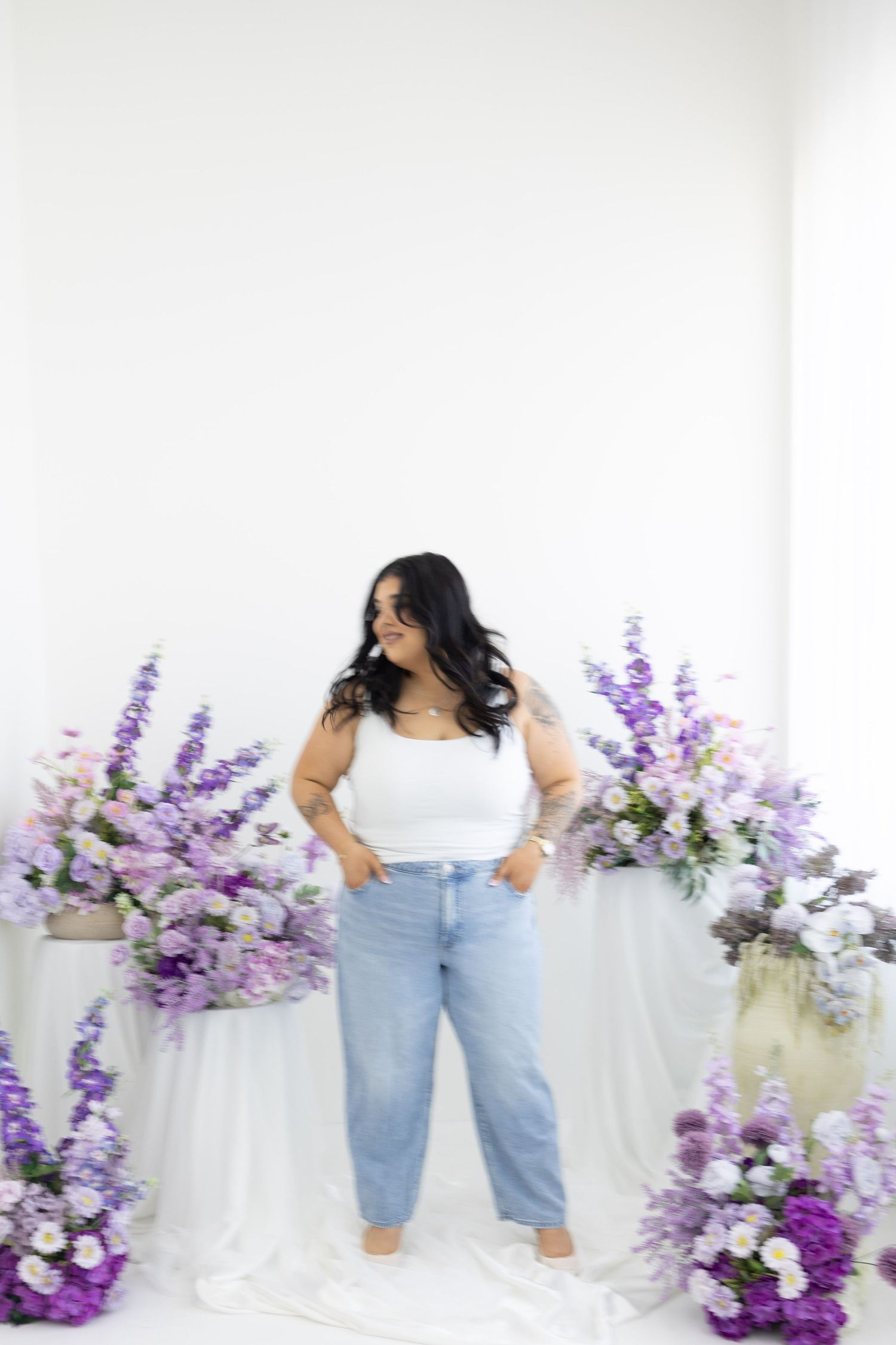 Woman in white crop top and light wash jeans posing among purple floral arrangements in studio.