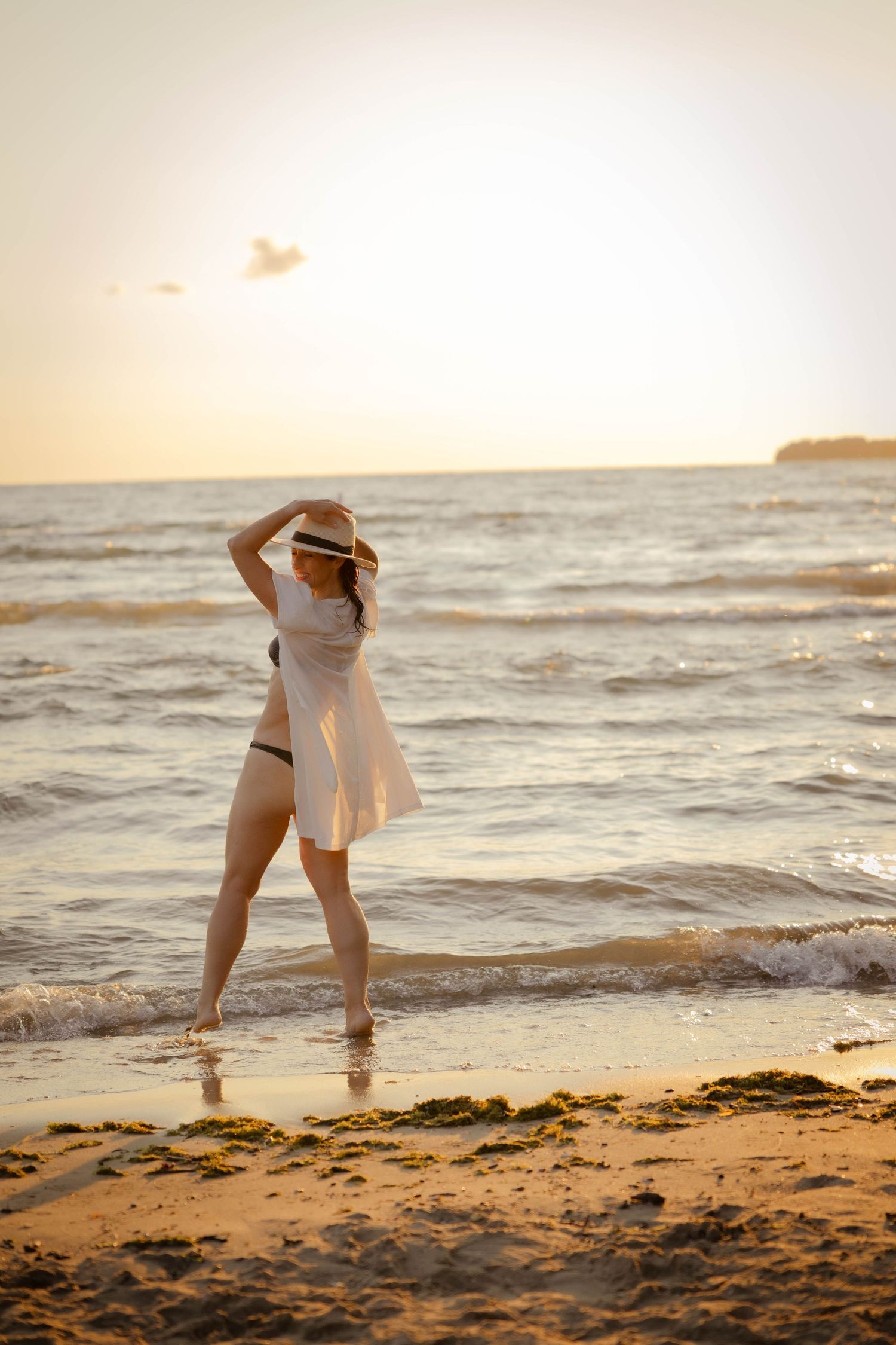 A person in a white sundress and hat stands at the edge of the ocean waves during a golden sunset.