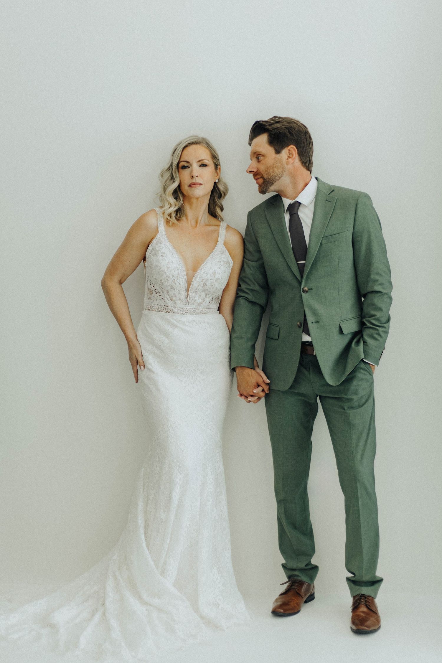 A bride in a white lace gown and groom in a green suit pose against a white wall in three artistic shots.