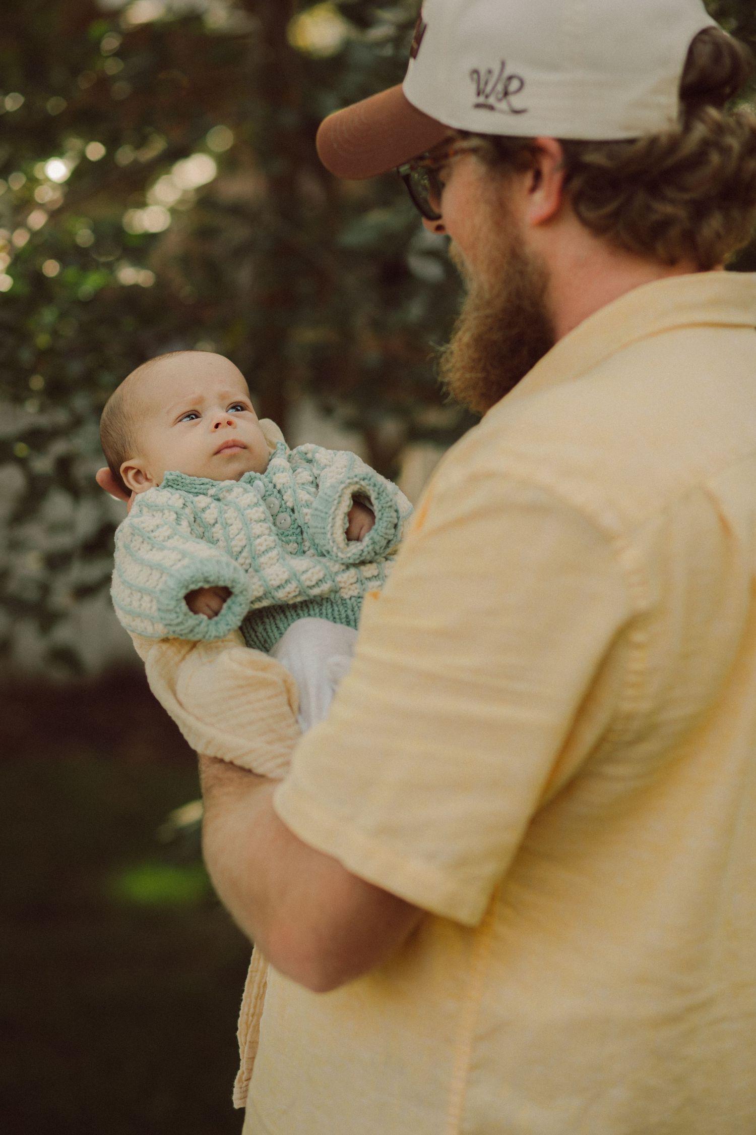 Bearded man in yellow shirt and cap holds a baby in a green knit sweater outdoors.