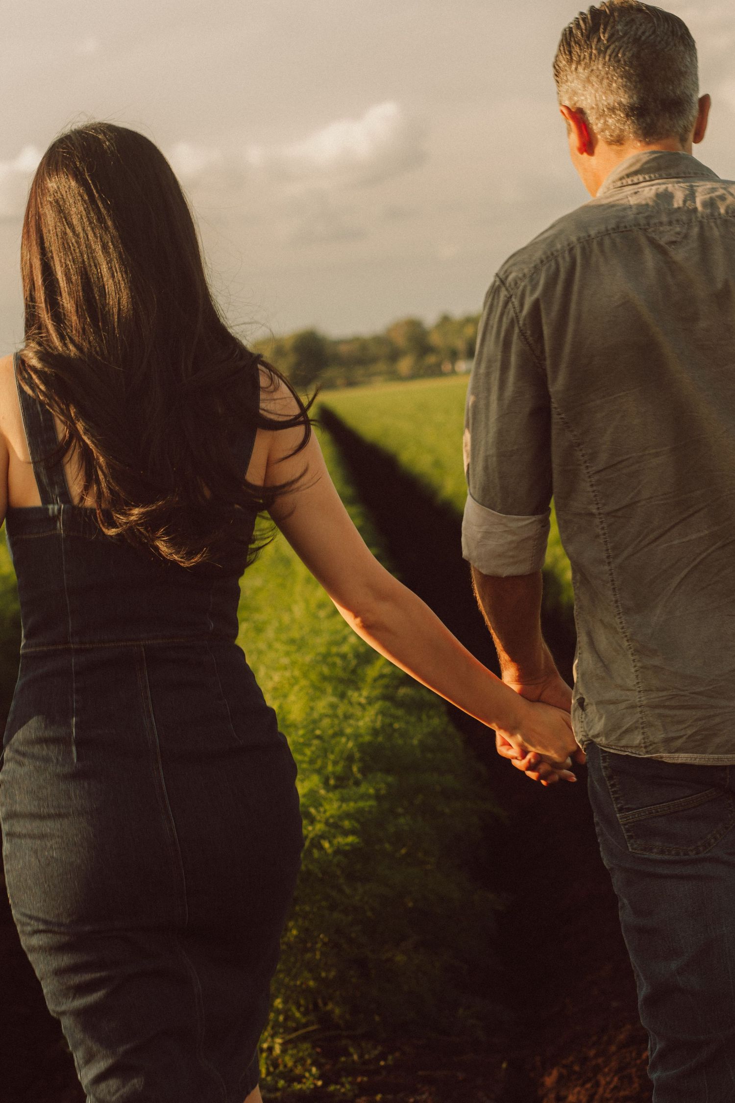 Couple holding hands walking through a lush green field at golden hour, viewed from behind.