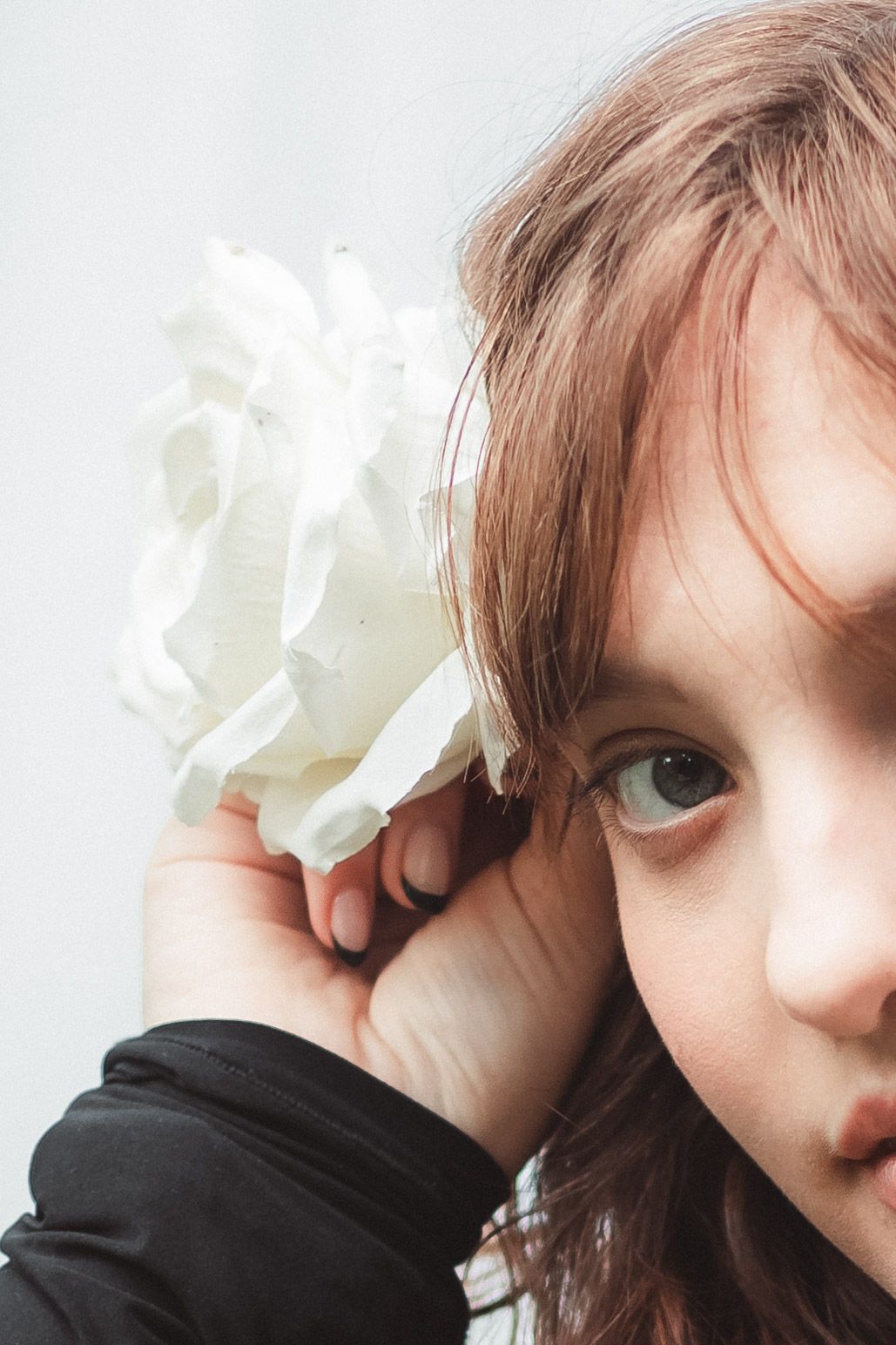 Young girl with brown hair holding a white flower accessory near her face against a light background.