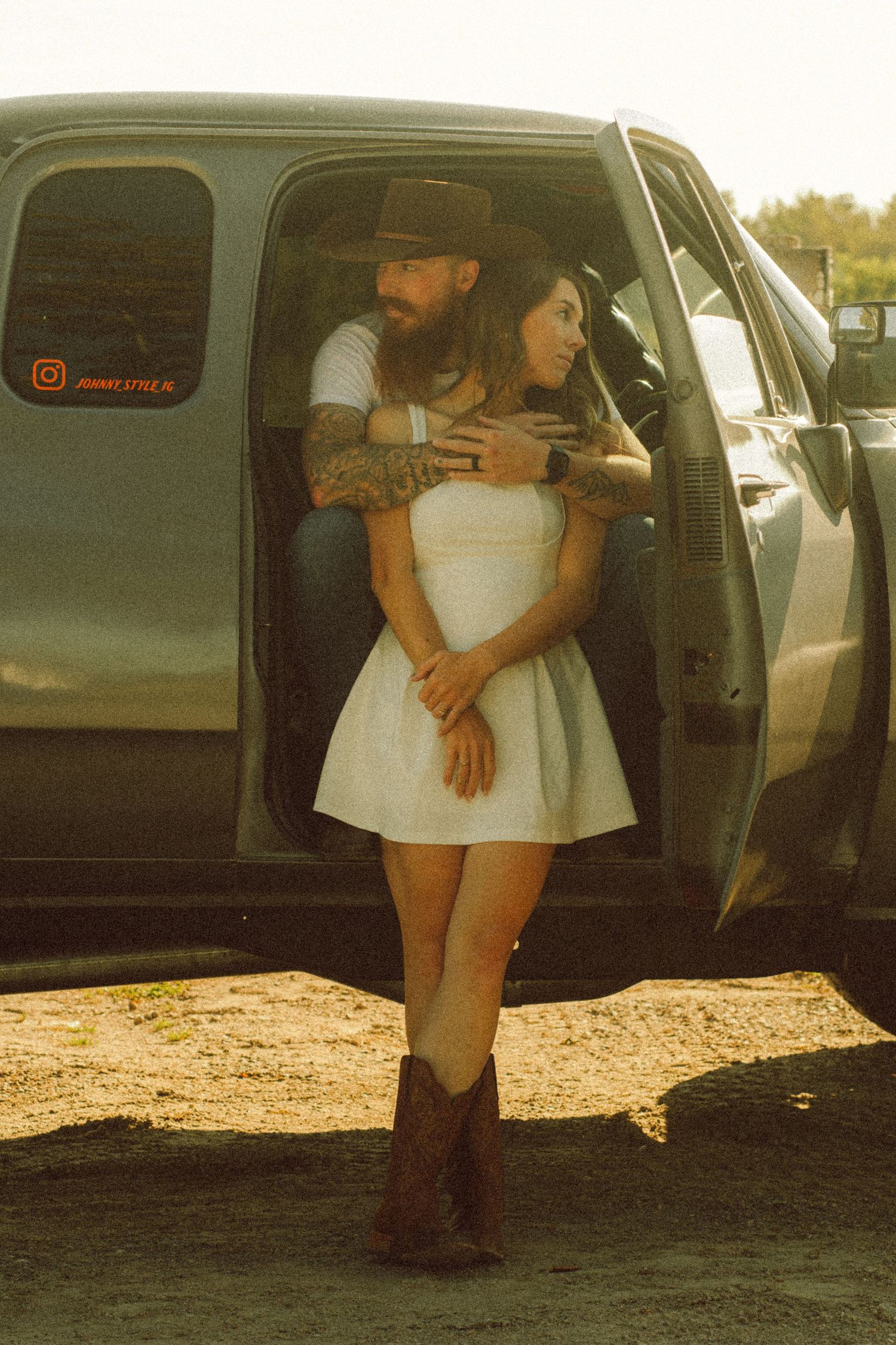 Couple embracing by a truck, woman in white dress and cowboy boots, man with tattoos and brown hat.