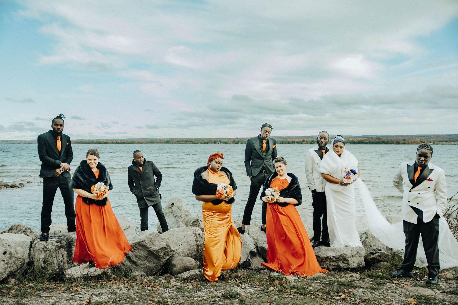 Wedding party poses playfully on rocky beach shoreline wearing orange and black formal attire.